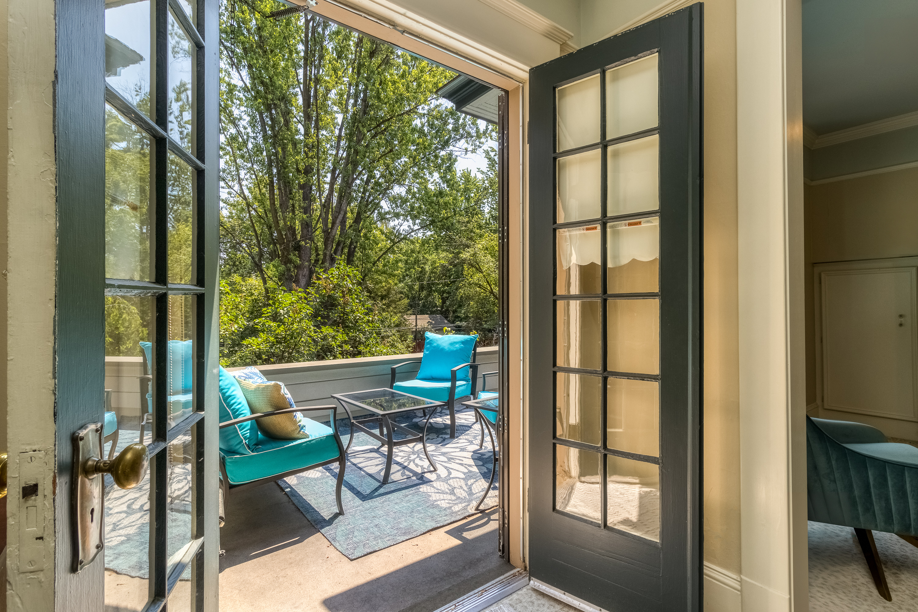 A set of French doors stand open revealing a sunny private balcony with cushioned chairs and a coffee table and views of lush green trees.
