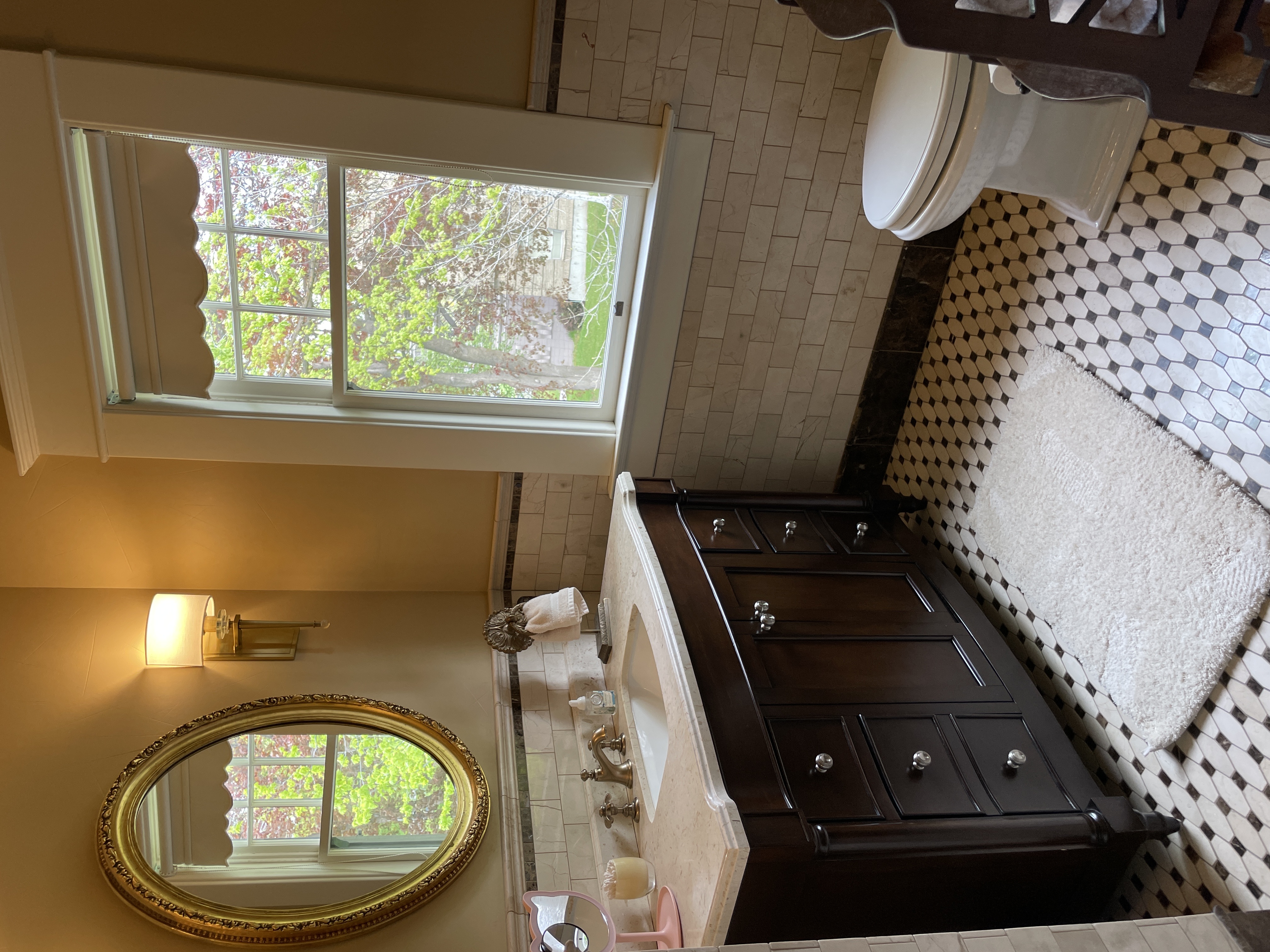 A classic bathroom featuring a dark wood vanity with a marble top, an oval gold mirror, a tiled floor, and a window.
