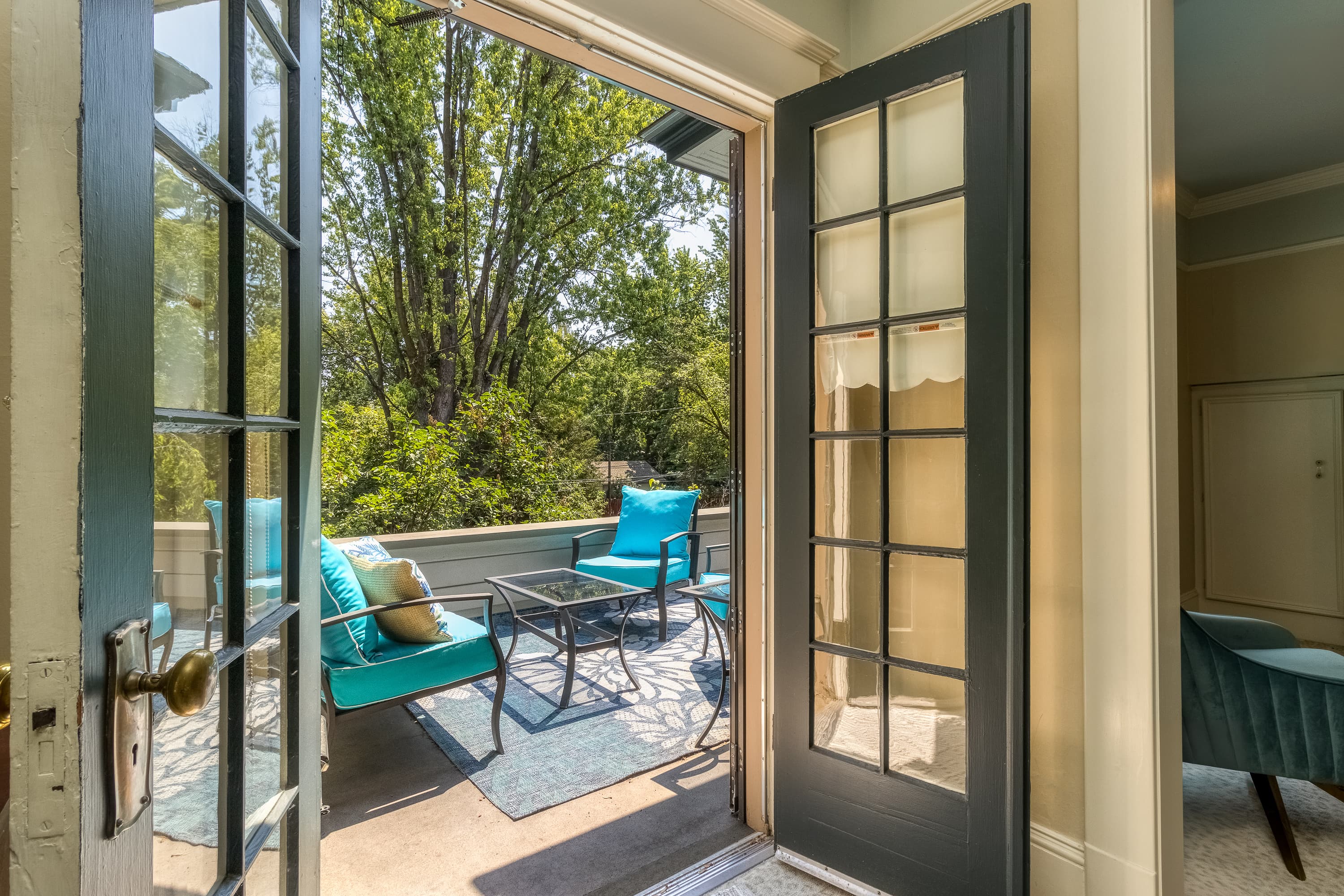 A set of French doors stand open revealing a sunny private balcony with cushioned chairs and a coffee table and views of lush green trees.
