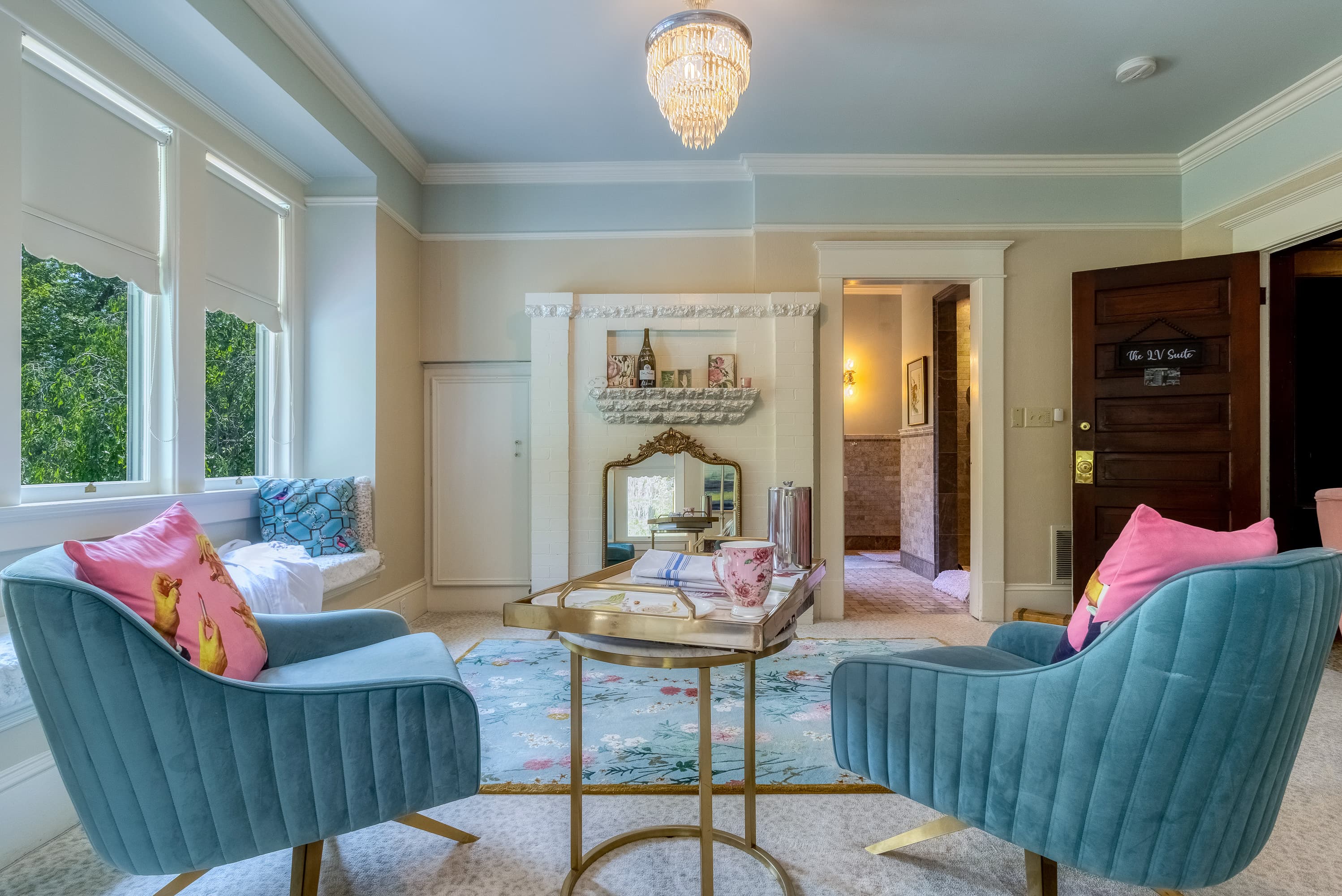 An elegantly decorated room with two teal armchairs featuring pink accent pillows, a small gold-framed coffee table, and a fireplace with a mirror above it.