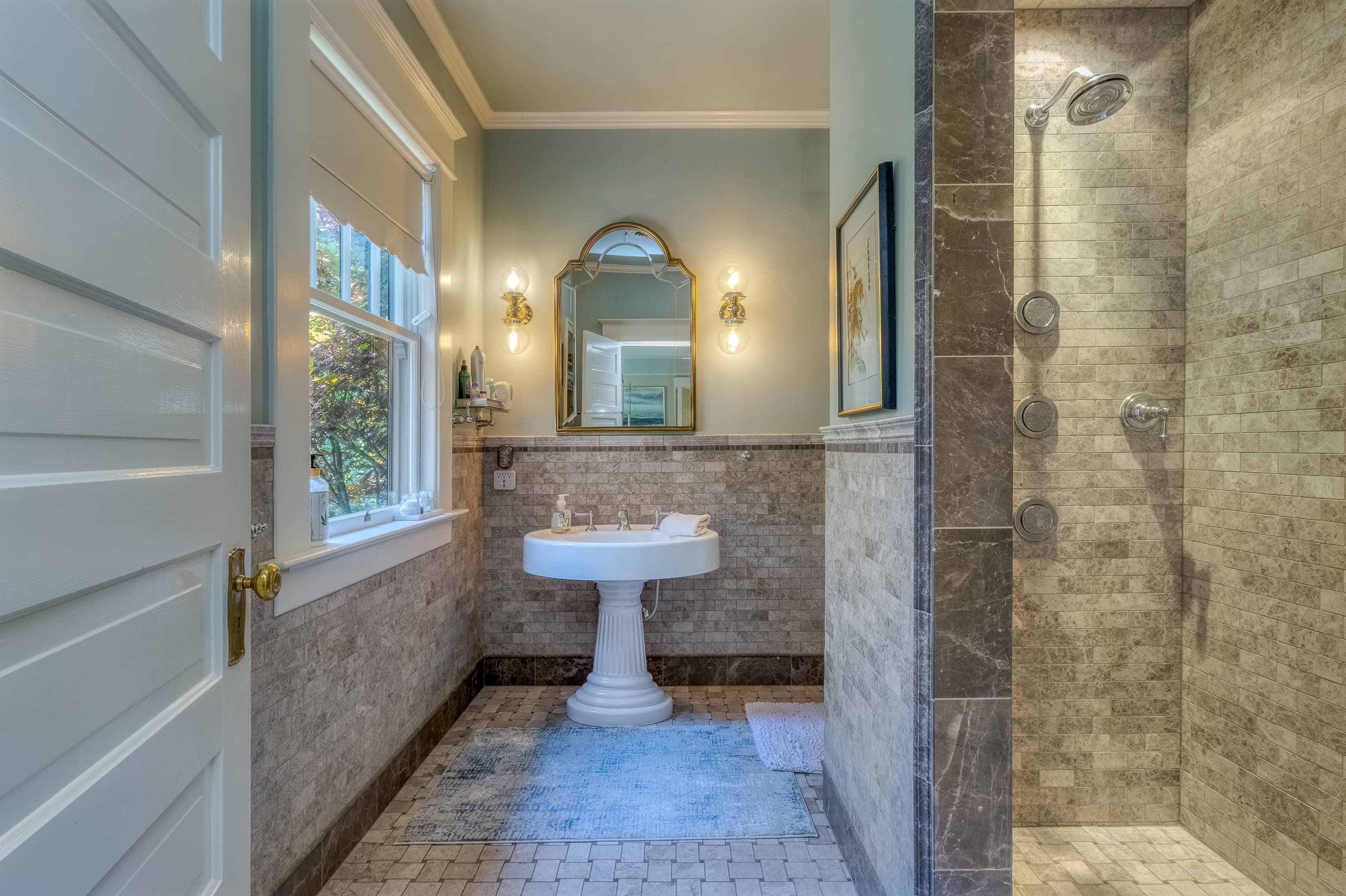 Elegant bathroom with a pedestal sink, gold mirror, and large walk-in stone shower.