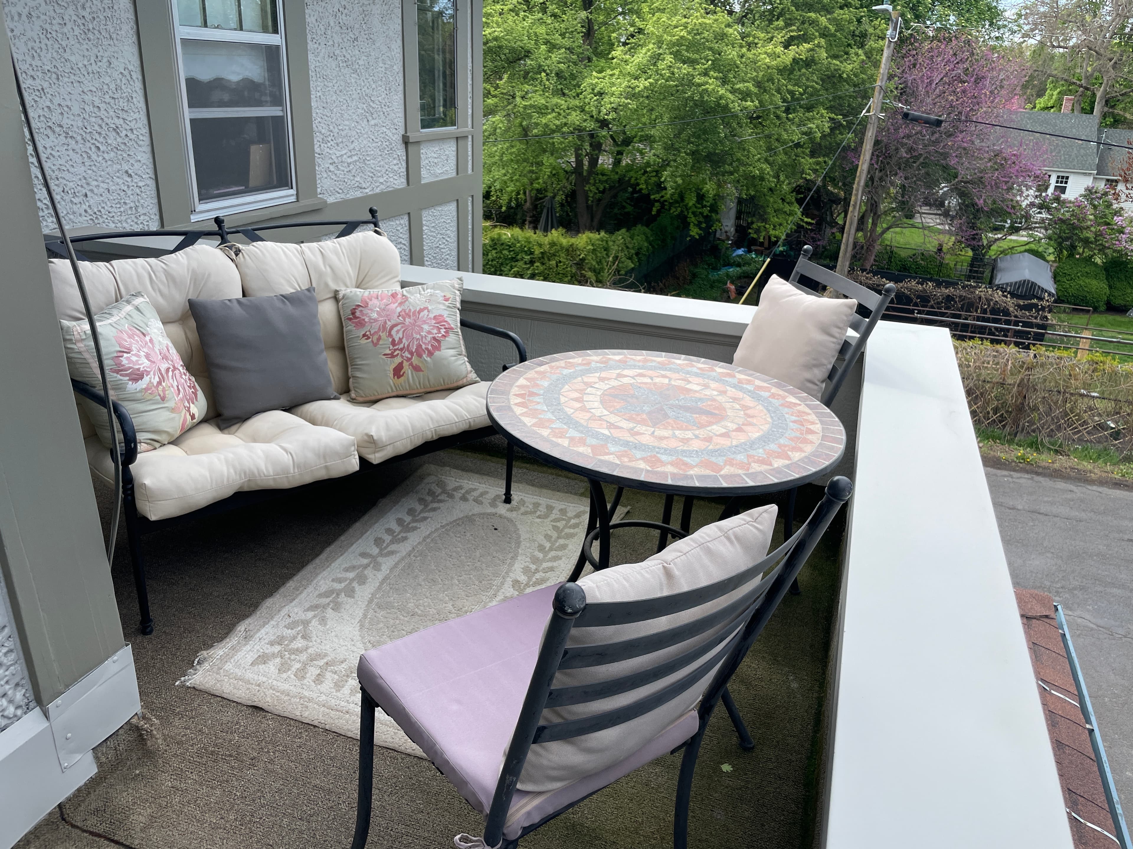 A pleasant outdoor patio with a round mosaic table, cushioned loveseat and chairs, set against a backdrop of lush greenery.
