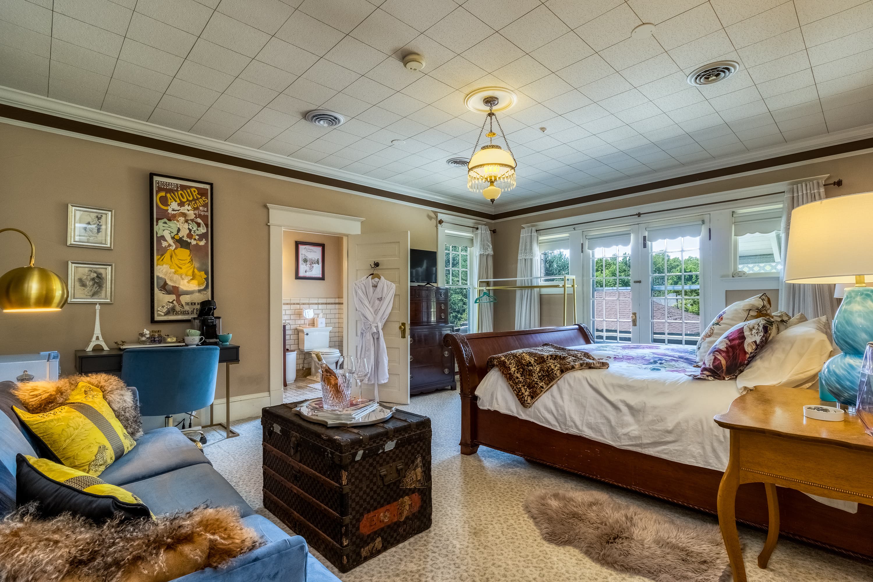 A spacious bedroom with a blue velvet sofa, vintage trunk, king bed, and an adjoining bathroom.