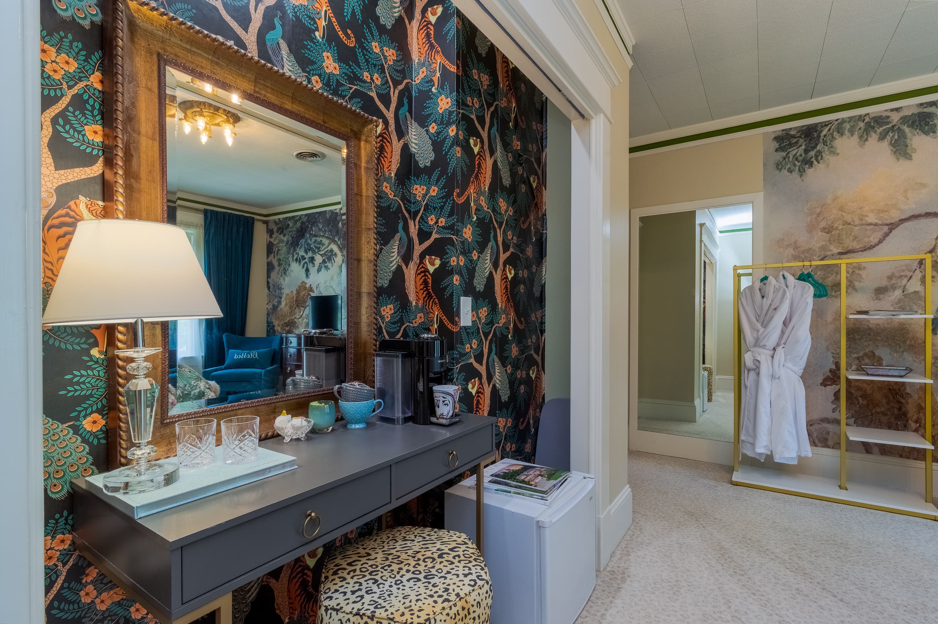 A lavish room featuring bold patterned wallpaper, a large antique mirror, a vanity table, and plush white bathrobes hanging on a clothing rack.