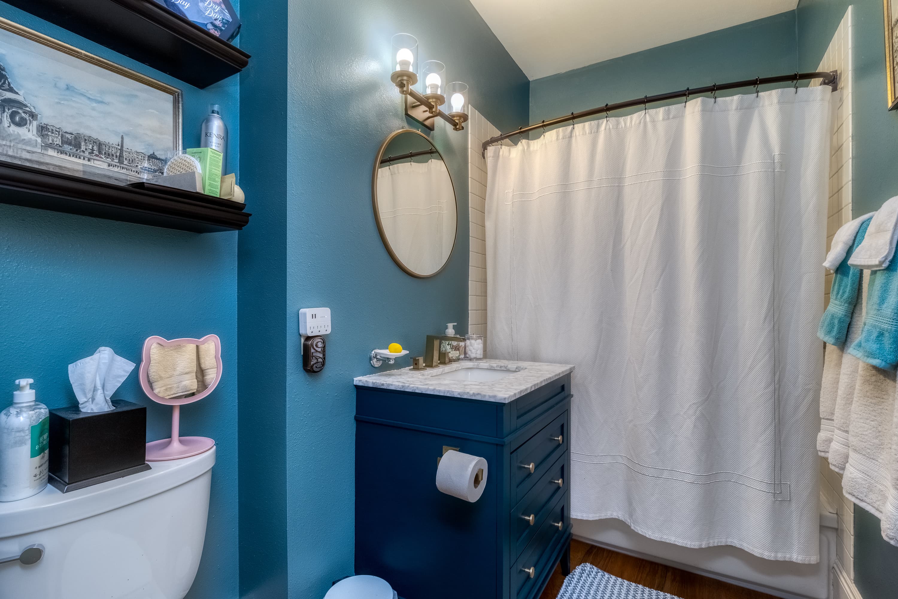 A small, vibrant bathroom with teal walls, a navy vanity, a round mirror, and a shower..