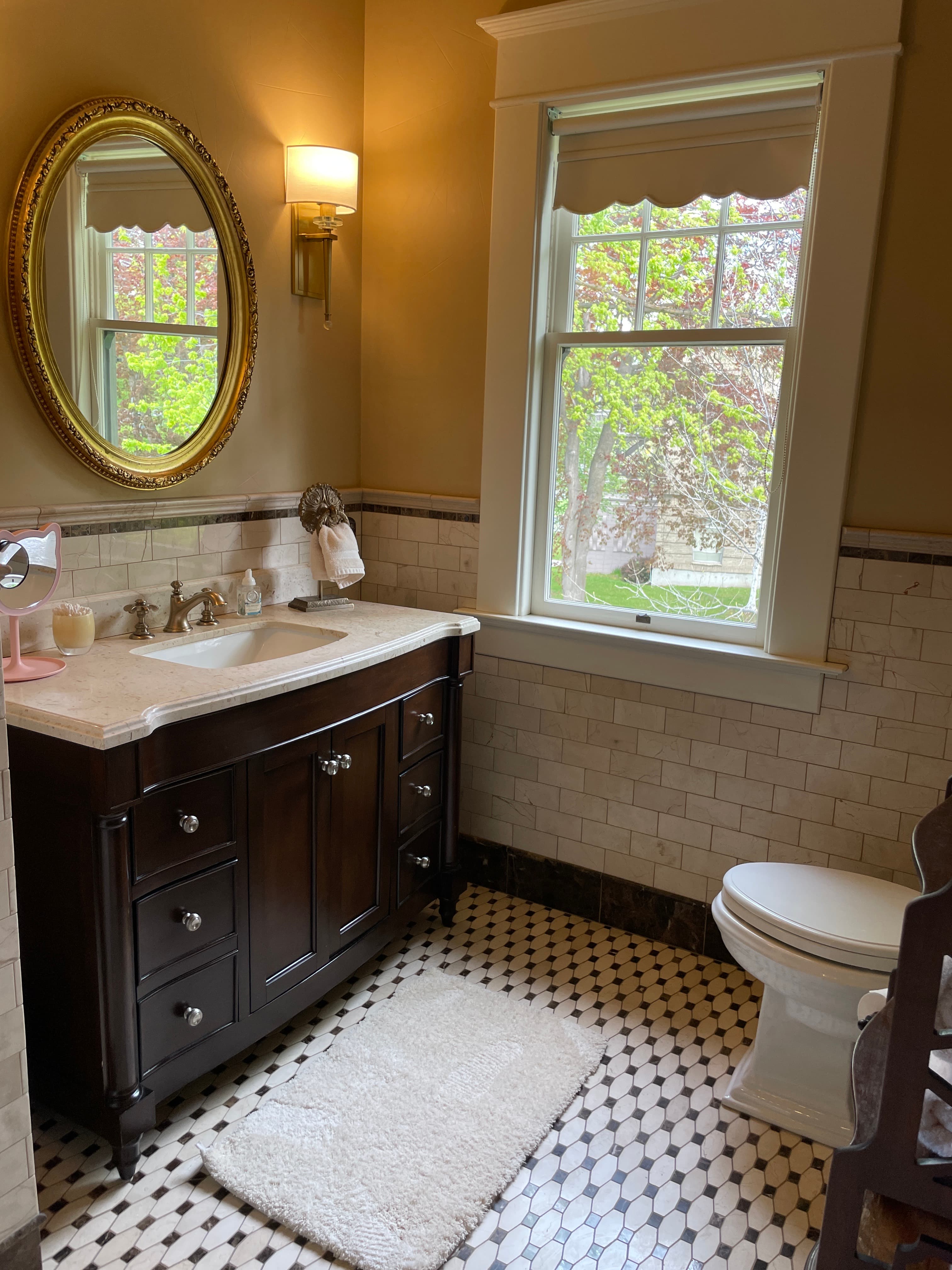 A classic bathroom featuring a dark wood vanity with a marble top, an oval gold mirror, a tiled floor, and a window.