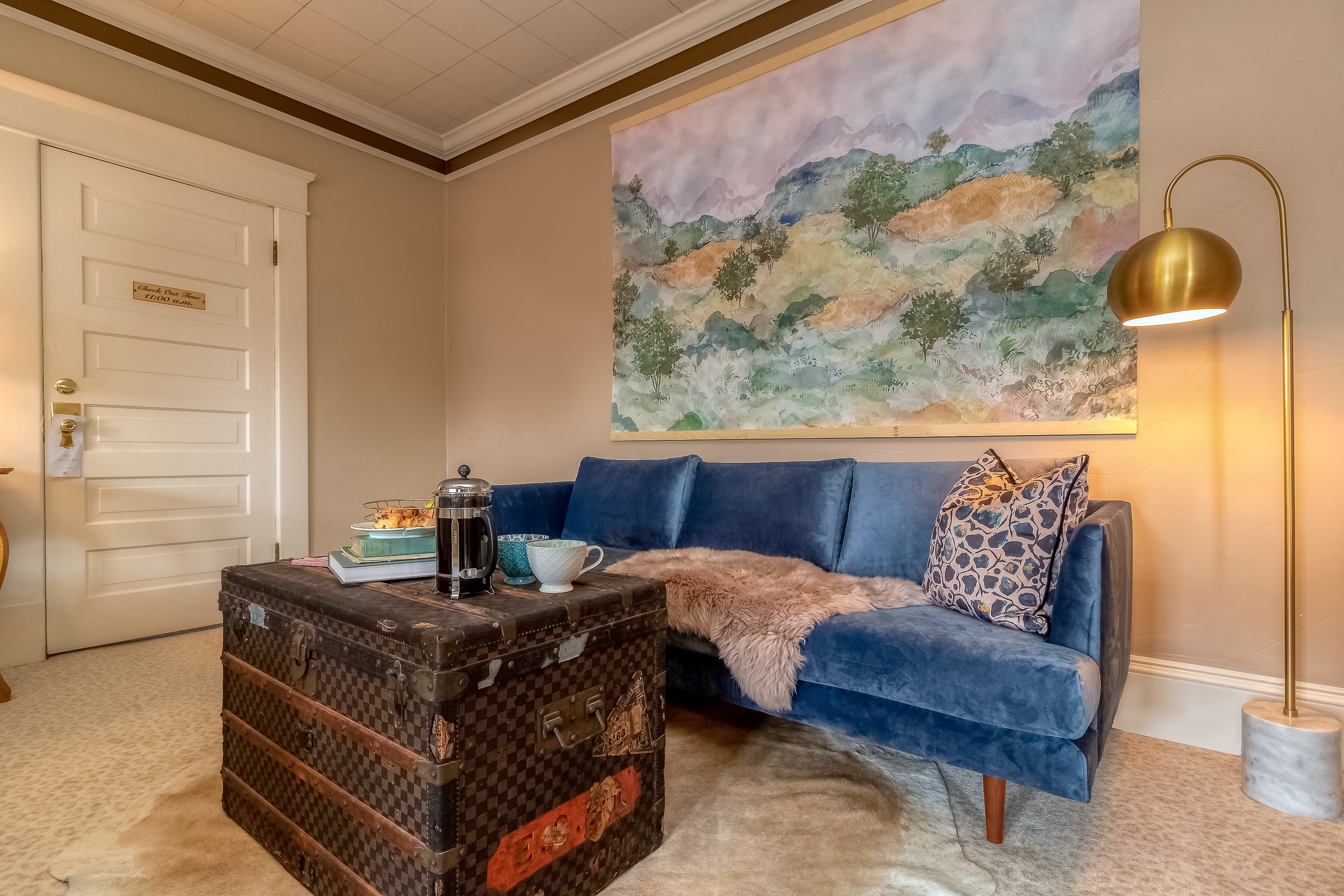 A chic sitting area featuring a blue velvet sofa, a vintage trunk coffee table, and a large landscape painting.