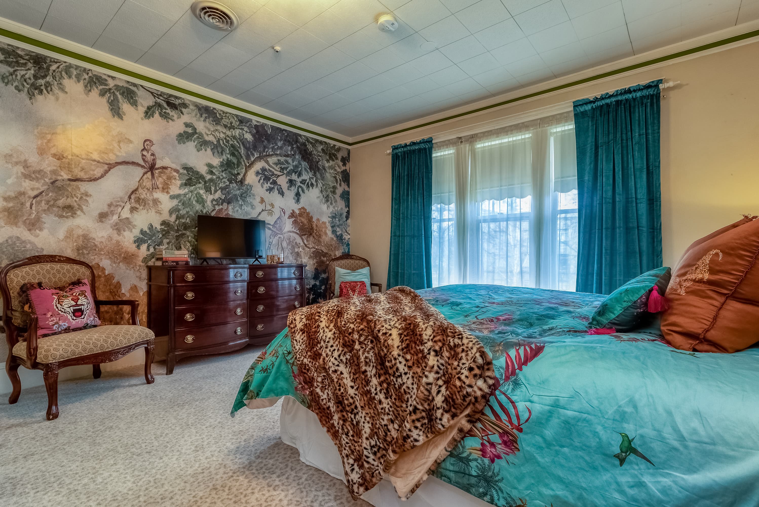 A richly decorated bedroom with an ornate mural wall, a large bed with colorful bedding, and vibrant turquoise curtains.