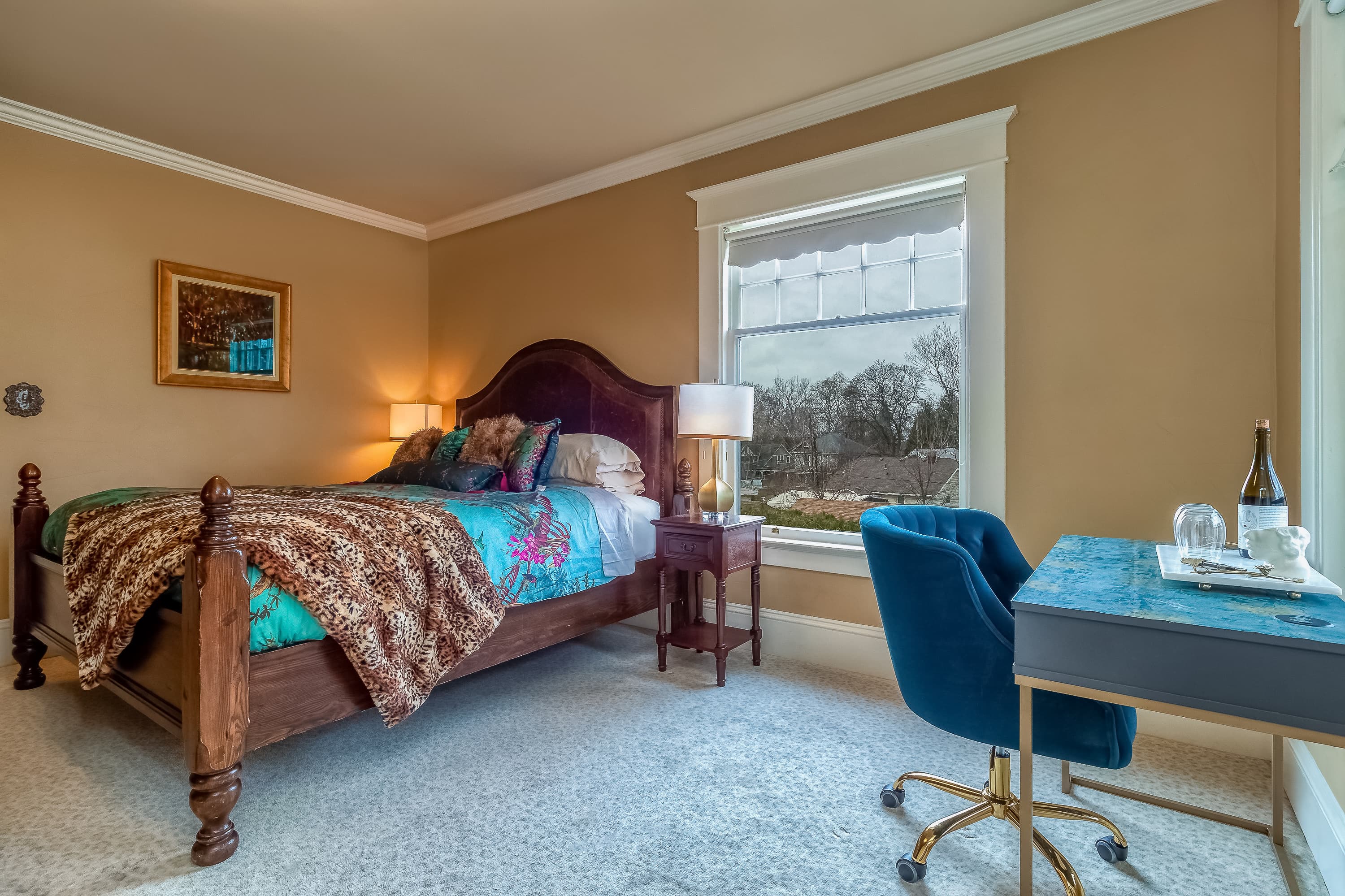 A warm bedroom with a wooden king-size bed, colorful bedding, and a stylish blue desk chair near a window, exuding comfort.