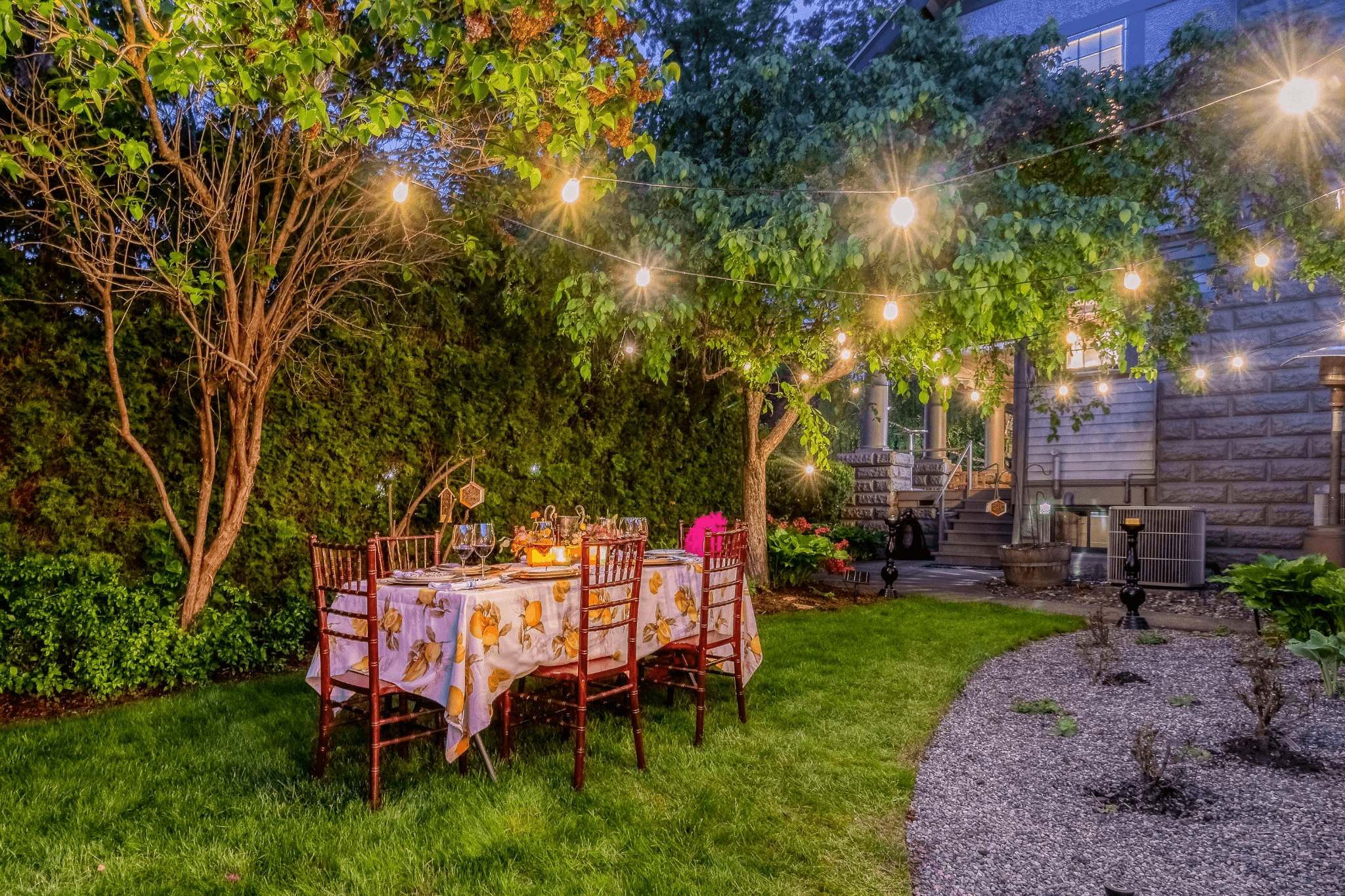 A beautifully set outdoor dining table illuminated by string lights under a tree in a lush garden.
