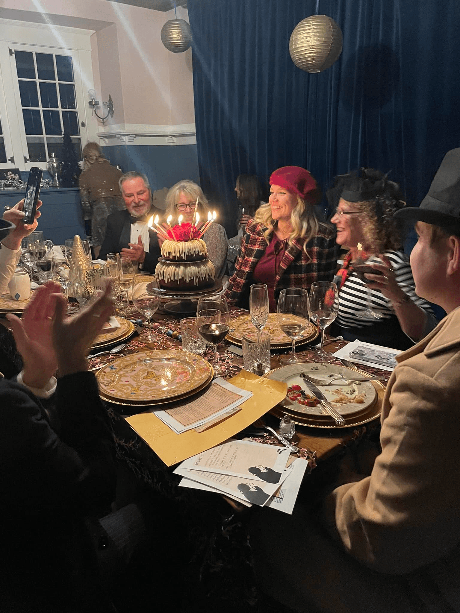A group of people gathers around a decorated table, celebrating with a birthday cake featuring lit candles.