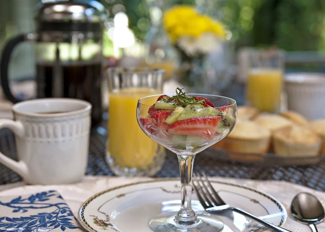 A glass dessert dish with mixed fruit sits on a beautifully set table with coffee, orange juice, and muffins in the background.