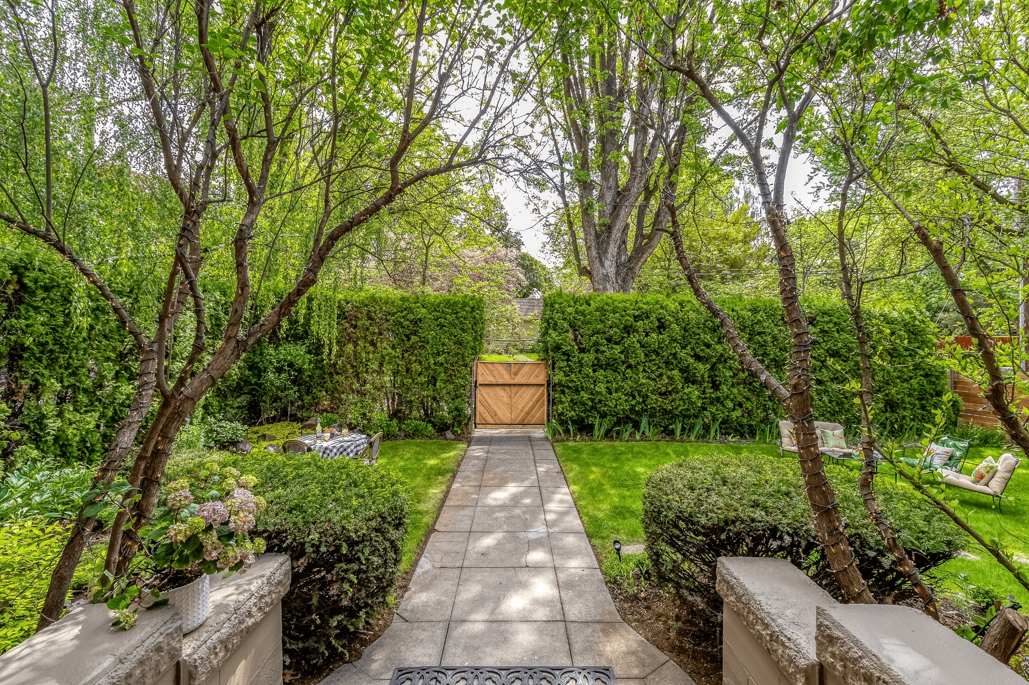 Stone path leading down from stone porch, through garden to wooden gate door set between tall green hedge