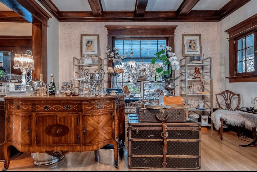 A cozy room featuring a wooden sideboard, vintage trunks, decorative plants, and elegant shelves filled with various ornaments.