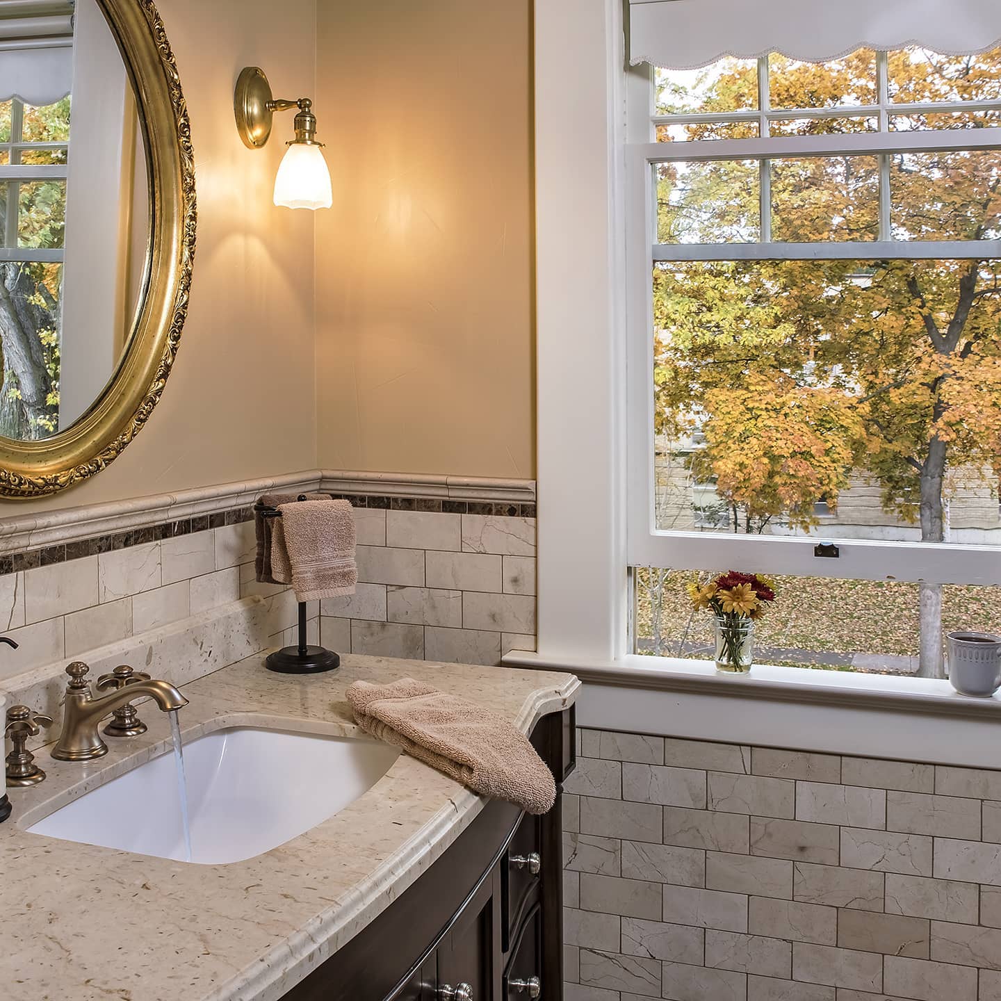 A stylish bathroom with a marble sink, decorative mirror, and a view of autumn leaves through a window.