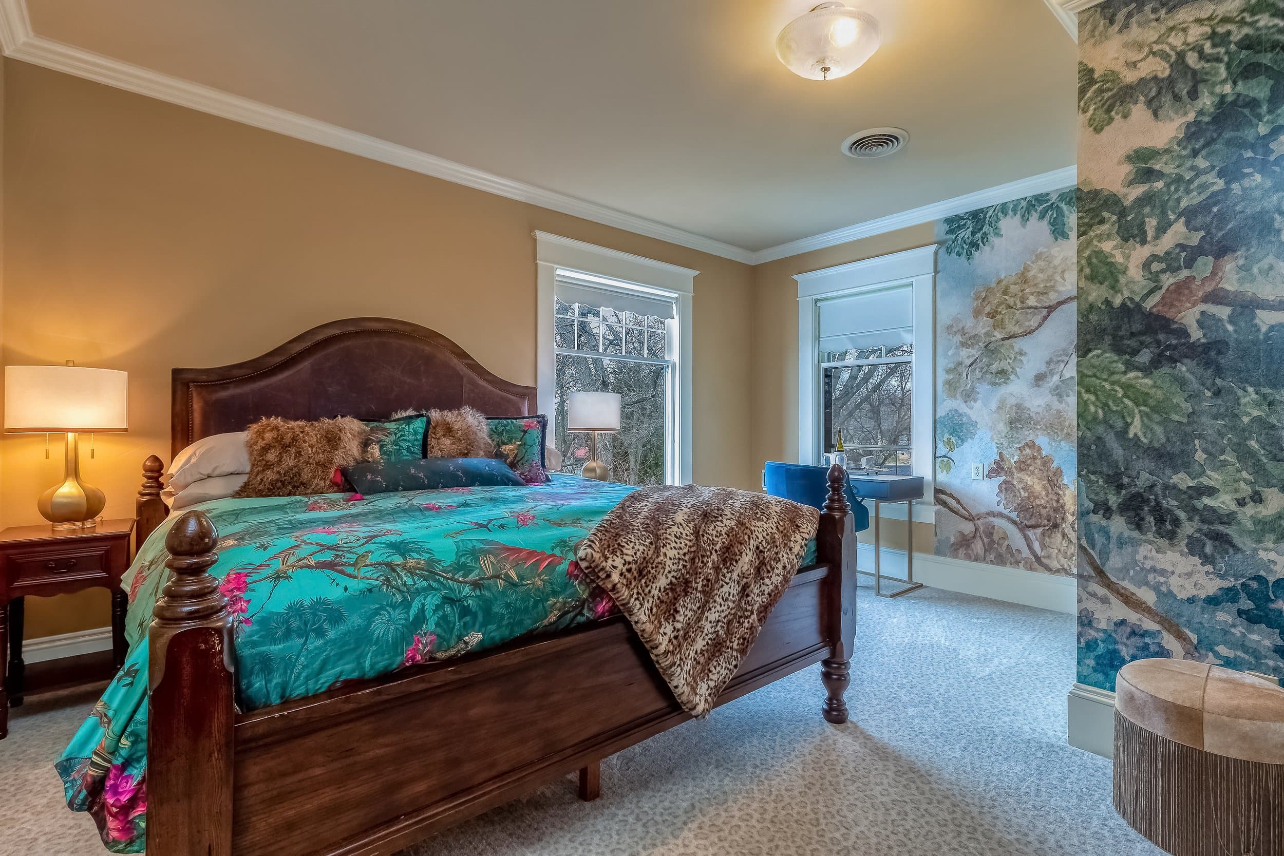 A cozy bedroom featuring a wooden bed with colorful bedding, a bedside lamp, and large windows.