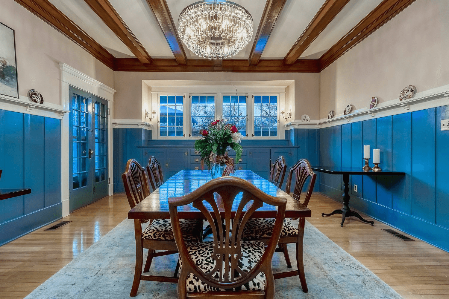 Shiny dark wood dining table atop a carpet in a blue-paneled dining room with a crystal chandelier hanging above