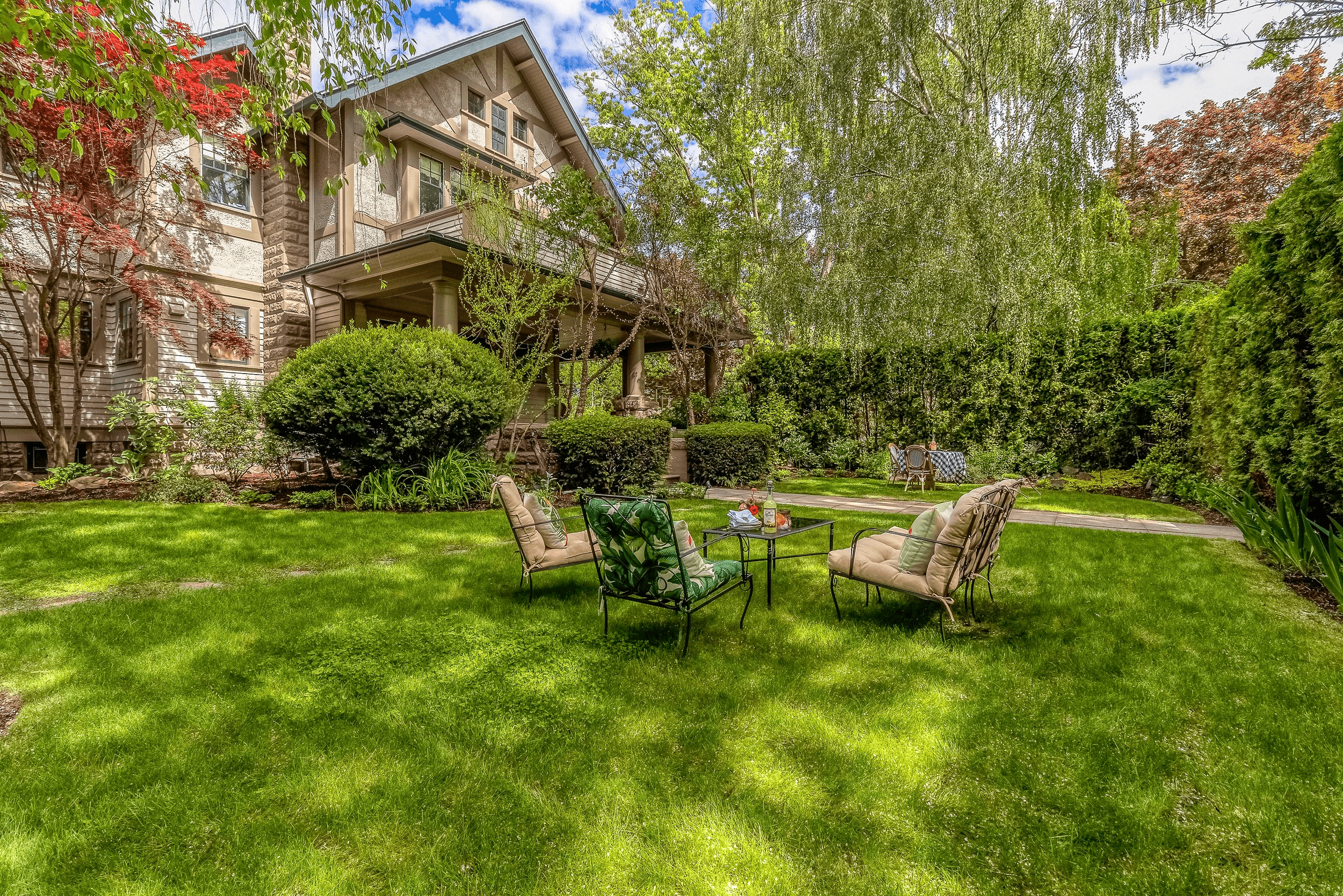 A lush green backyard featuring comfortable seating and a small table set amidst trees and shrubs.