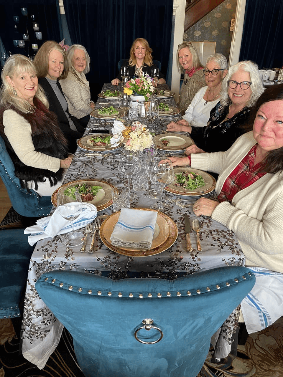 A group of eight women gathers around an elegantly set dining table adorned with salads and flowers.