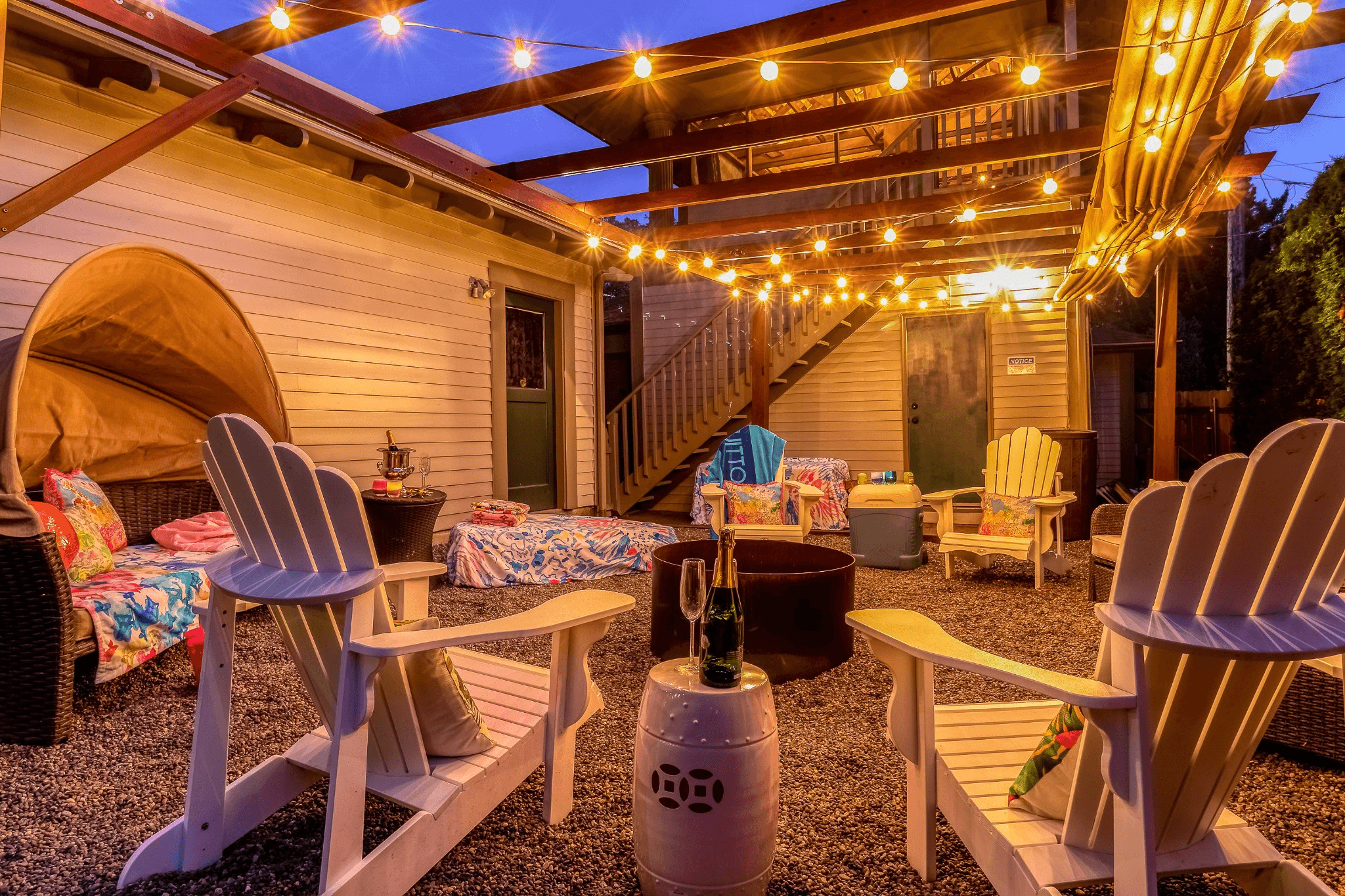 A cozy outdoor patio with string lights, seating, and a fire pit, surrounded by decorative elements.