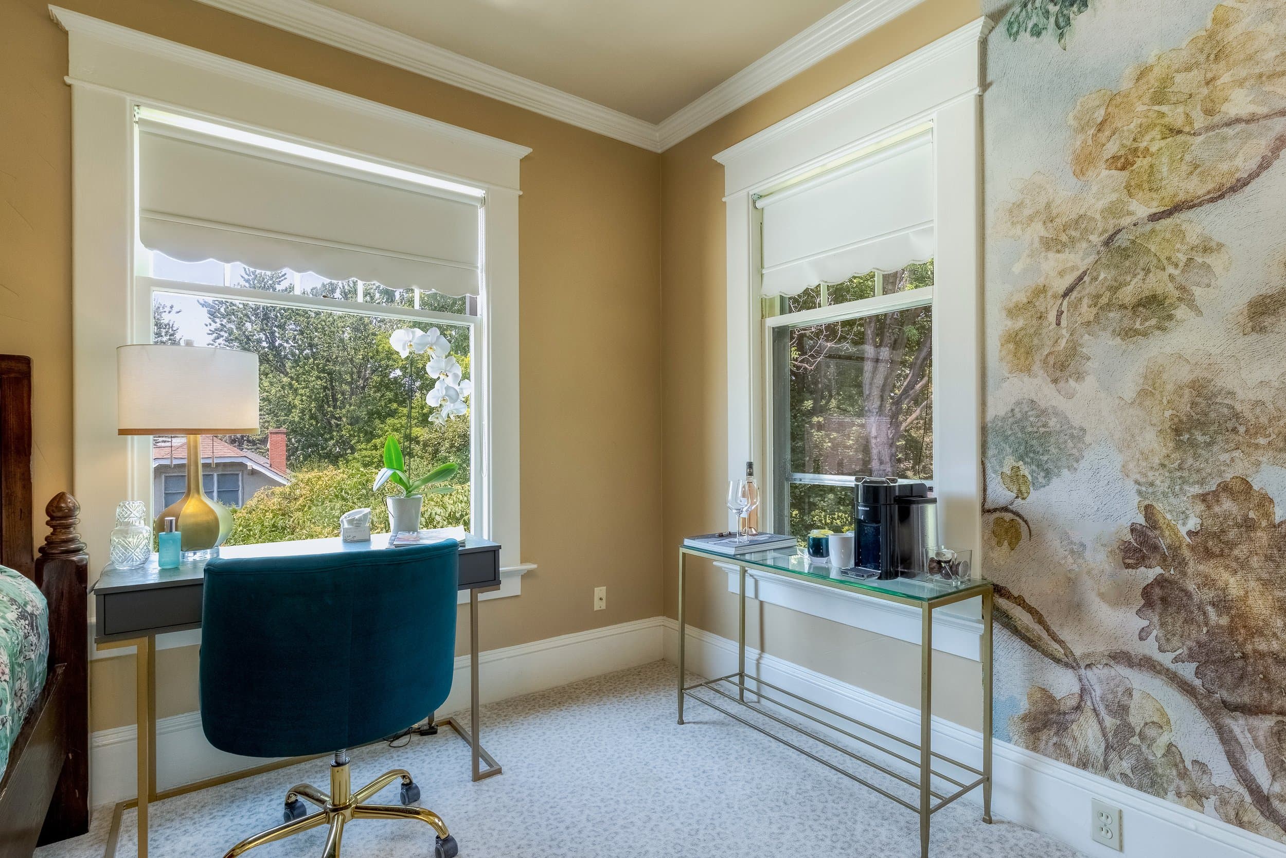 A cozy office space features a desk with a teal chair, two windows with views, and a decorative floral wall.