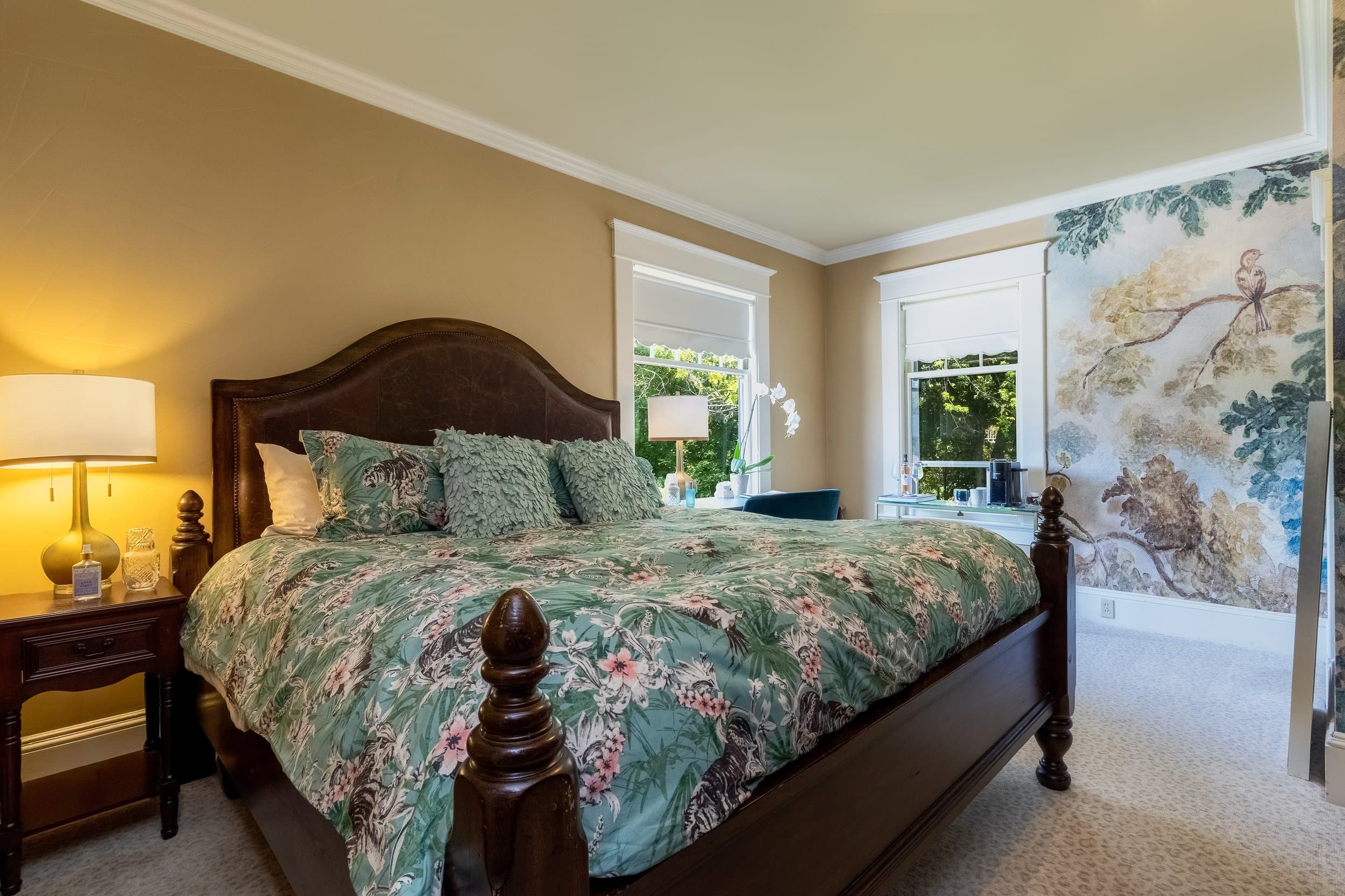 A cozy bedroom featuring a large bed with a floral comforter, bedside table with a lamp, and windows adorned with soft natural light.