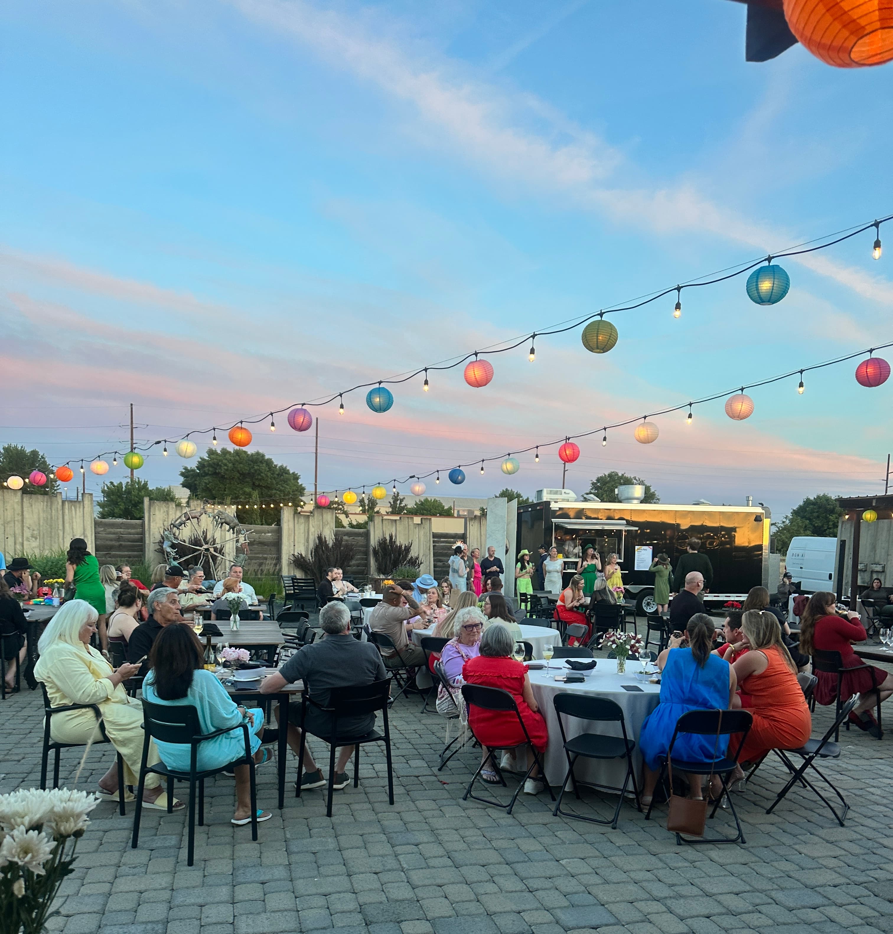 A lively outdoor gathering at sunset with people seated at tables under colorful lanterns. A lively outdoor gathering at sunset with people seated at tables under colorful lanterns.