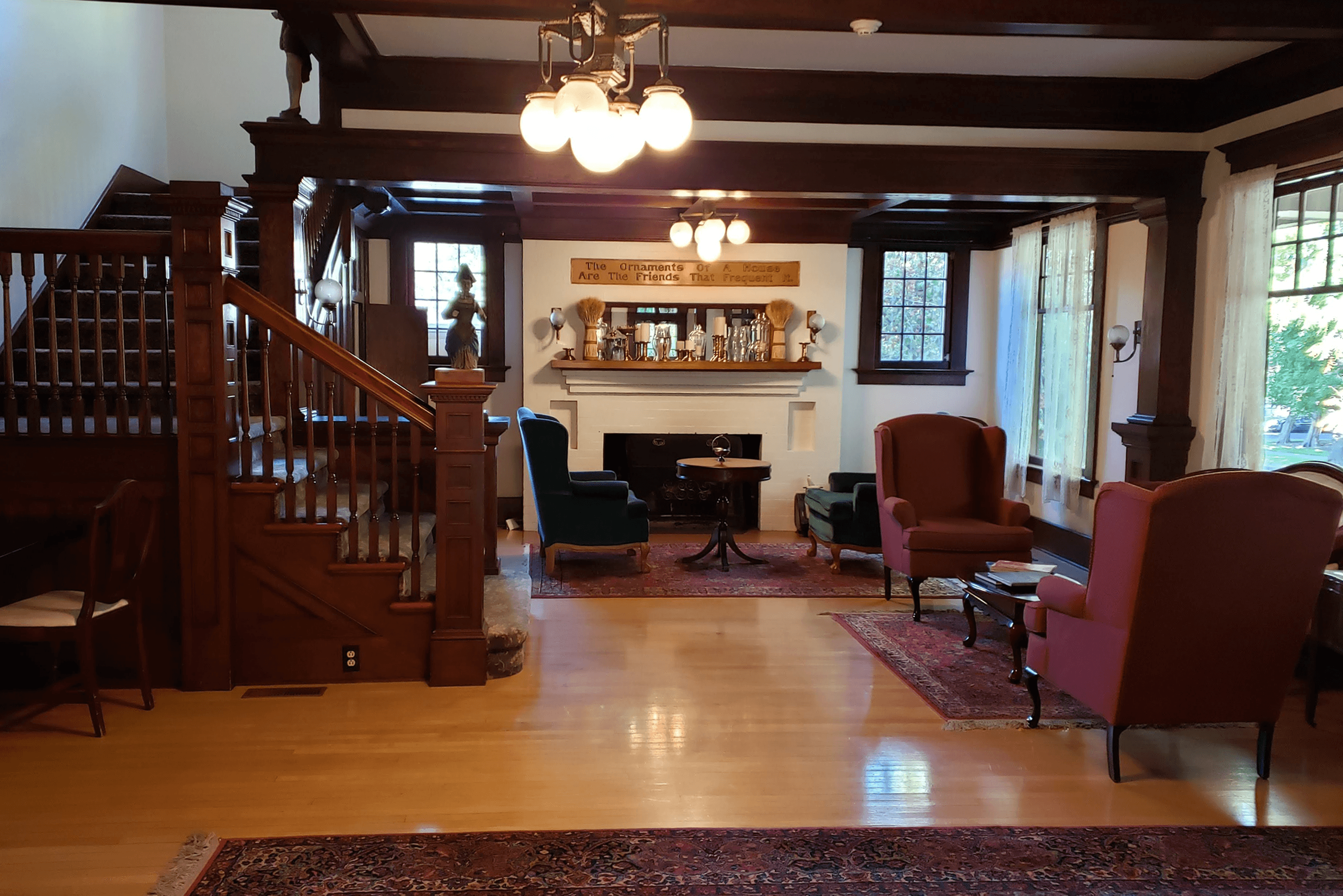 Seating area of a manor house with a dark staircase to the left and matching, wine-colored wing chairs to the right, a fireplace in the rear