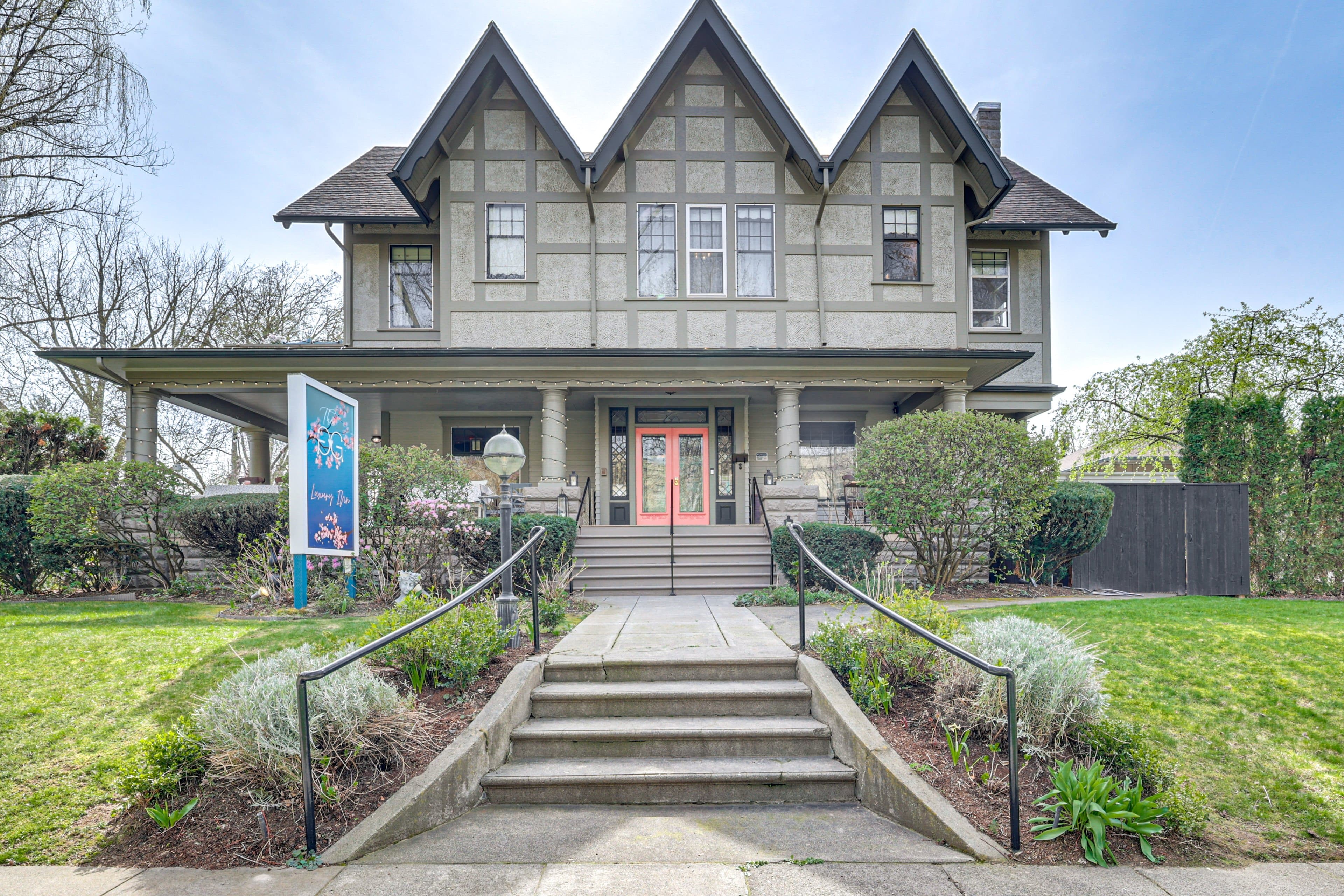 A large, two-story house with a gray exterior and decorative wooden trim, featuring a front staircase and landscaping.