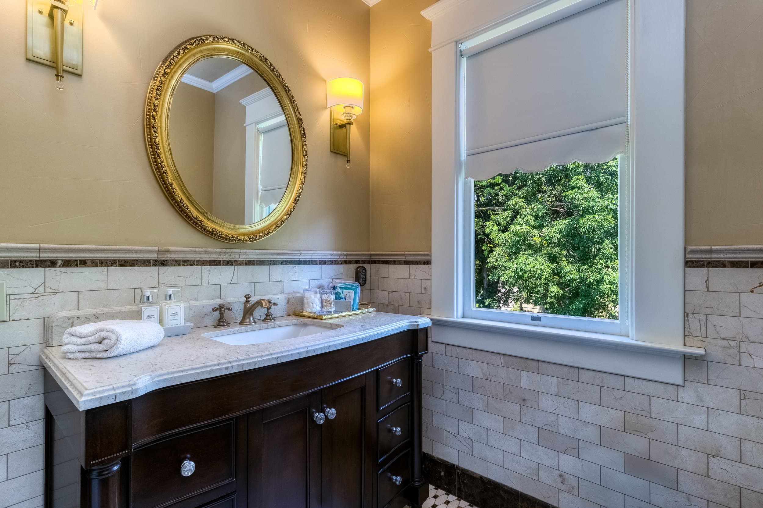 A stylish bathroom featuring a marble countertop, gold-framed mirror, and window overlooking greenery.