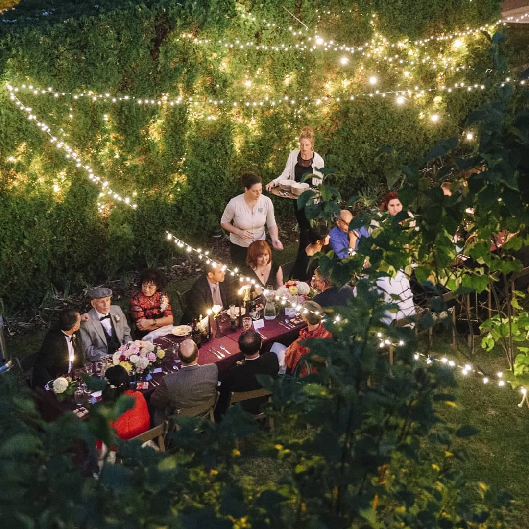A festive outdoor dinner party is illuminated by string lights with guests seated around a long table.