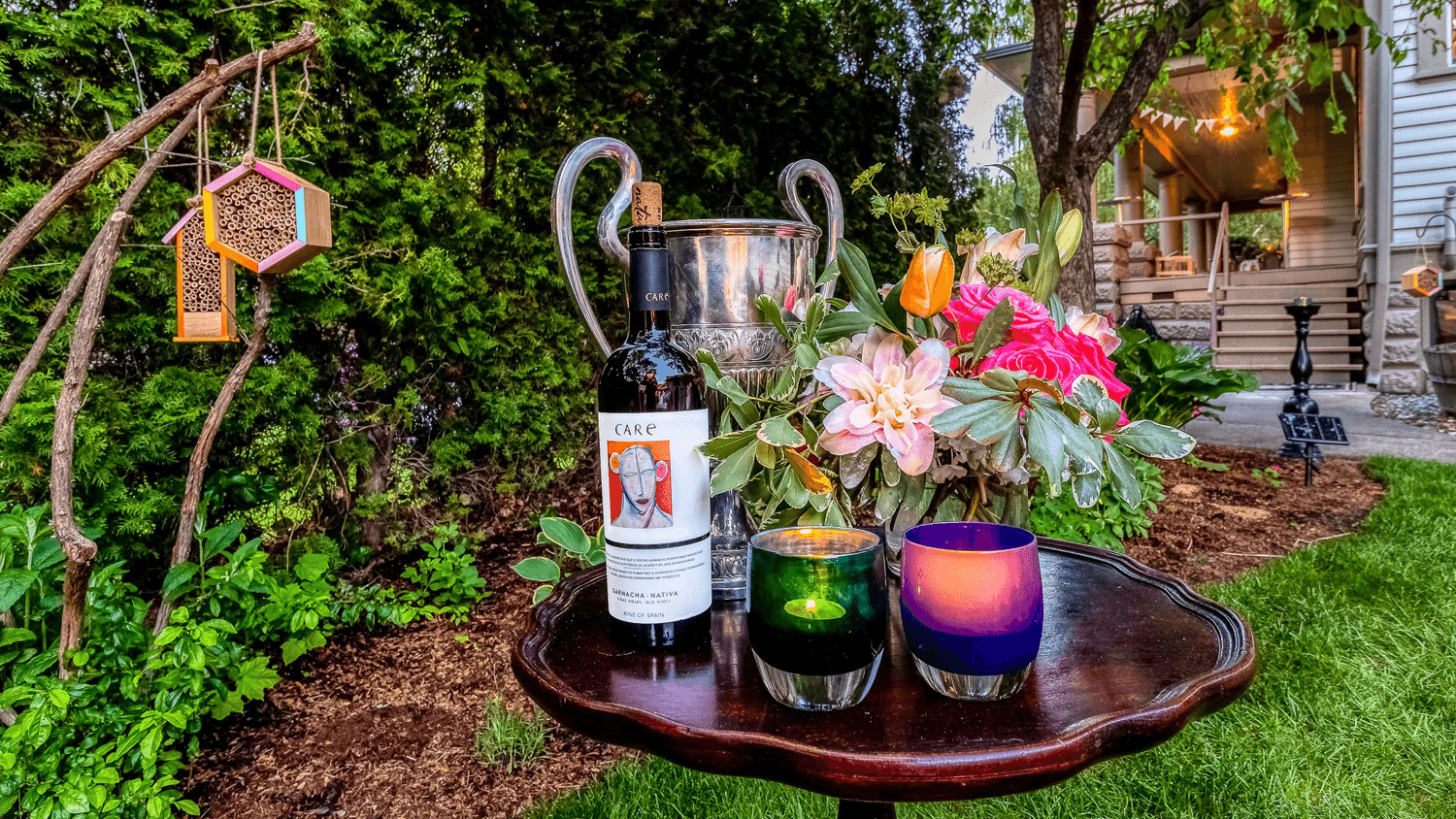 Silver ice bucket, bouquet of flowers, two colorful candles, and a bottle of wine sit on a small wooden side table, set outdoors in the garden