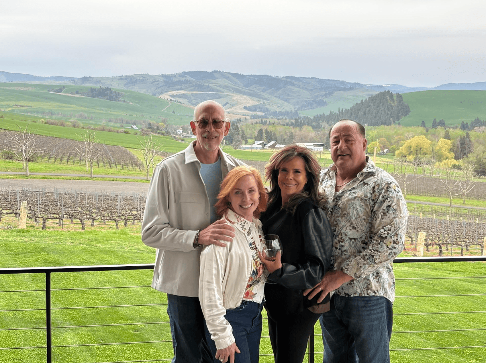 A group of four people pose together on a balcony with a scenic vineyard and rolling hills in the background.