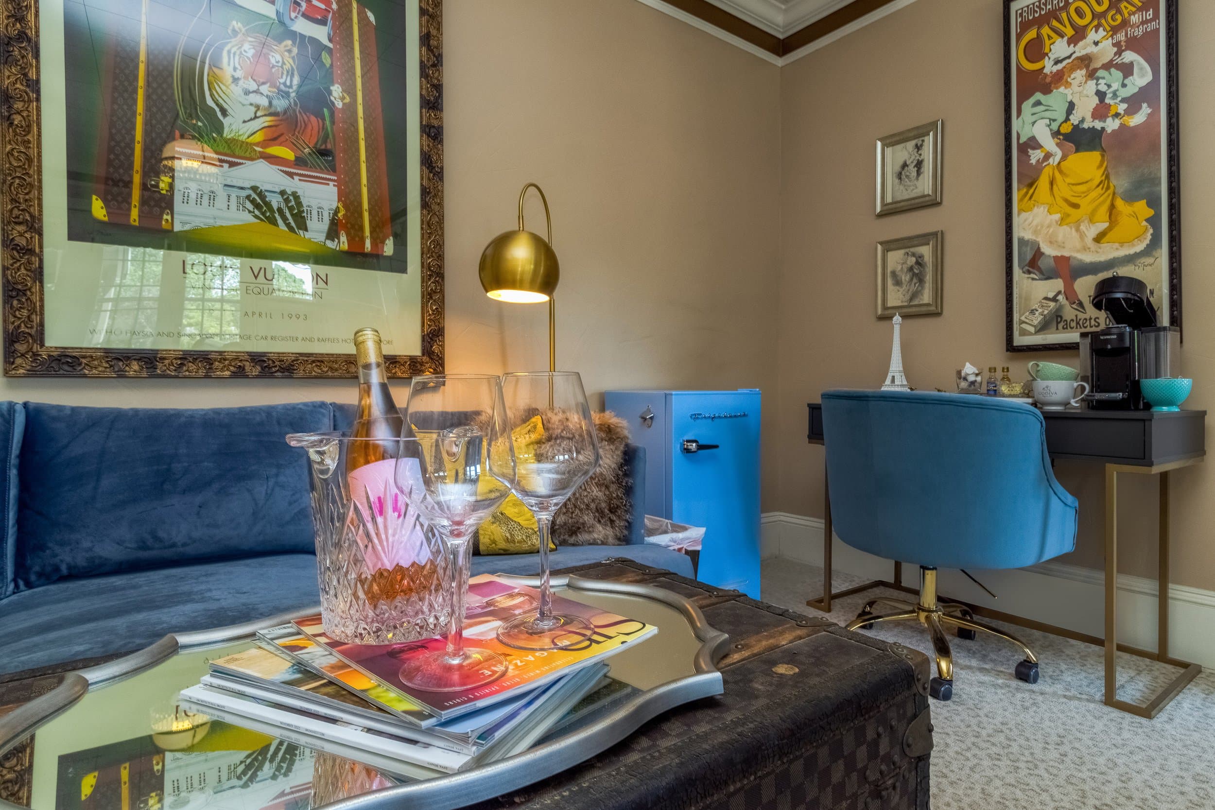 A stylish living space featuring a blue couch, a small blue refrigerator, a desk with a coffee maker, and decorative artwork on the walls.