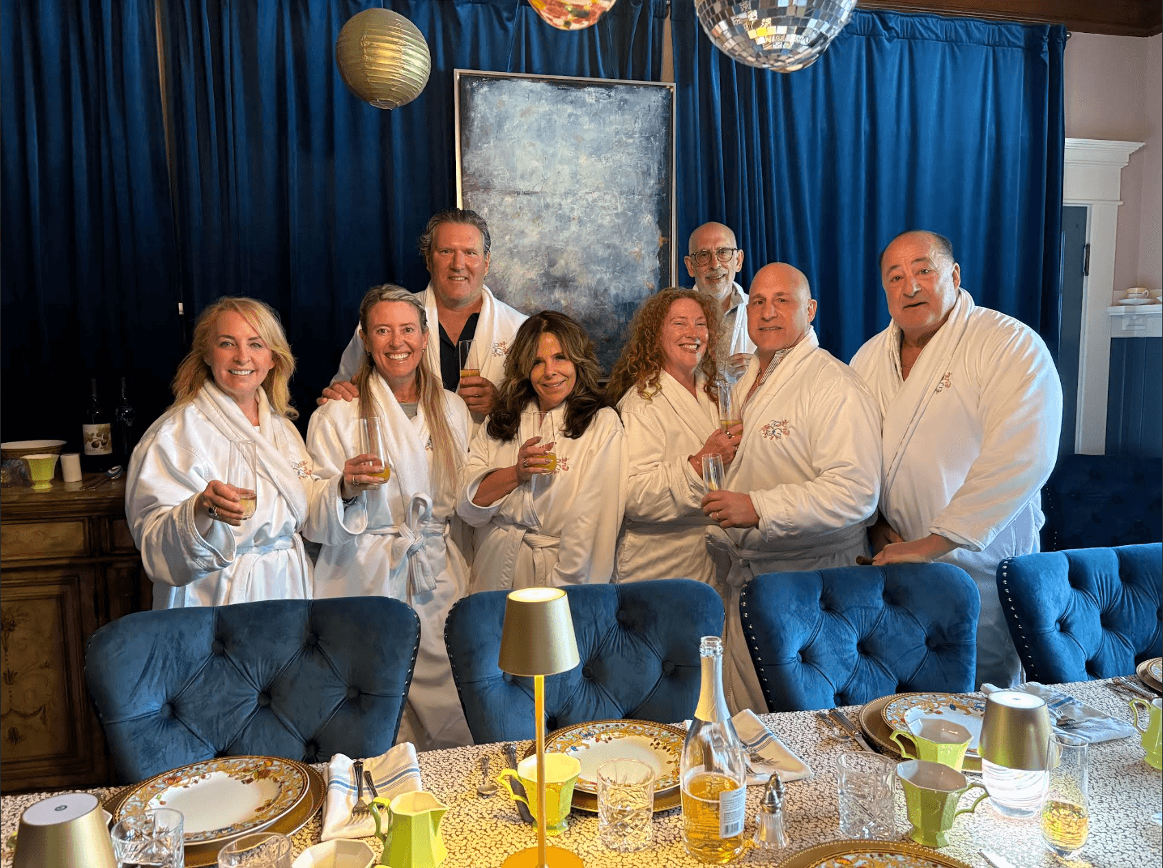 A group of eight people in white robes smile and toast with drinks in a cozy dining room setting.