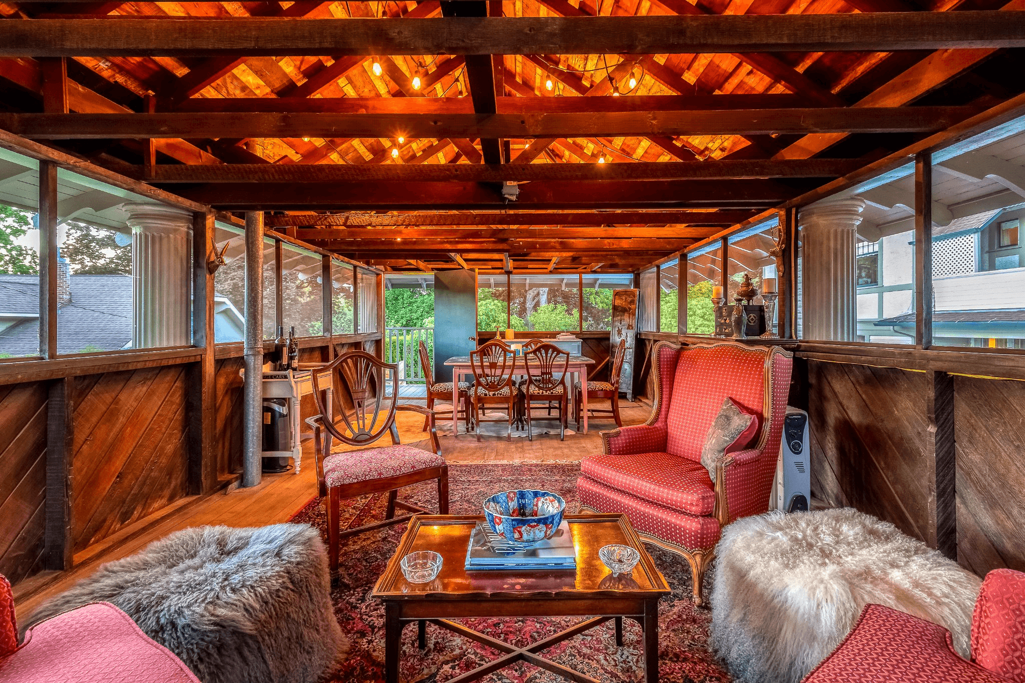 A cozy, well-lit indoor seating area features plush chairs, a wooden dining table, and large windows overlooking greenery.