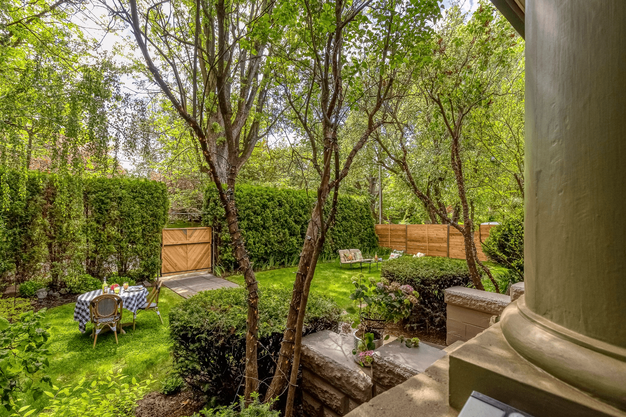 A lush garden featuring a wooden gate, patio furniture, and vibrant greenery.