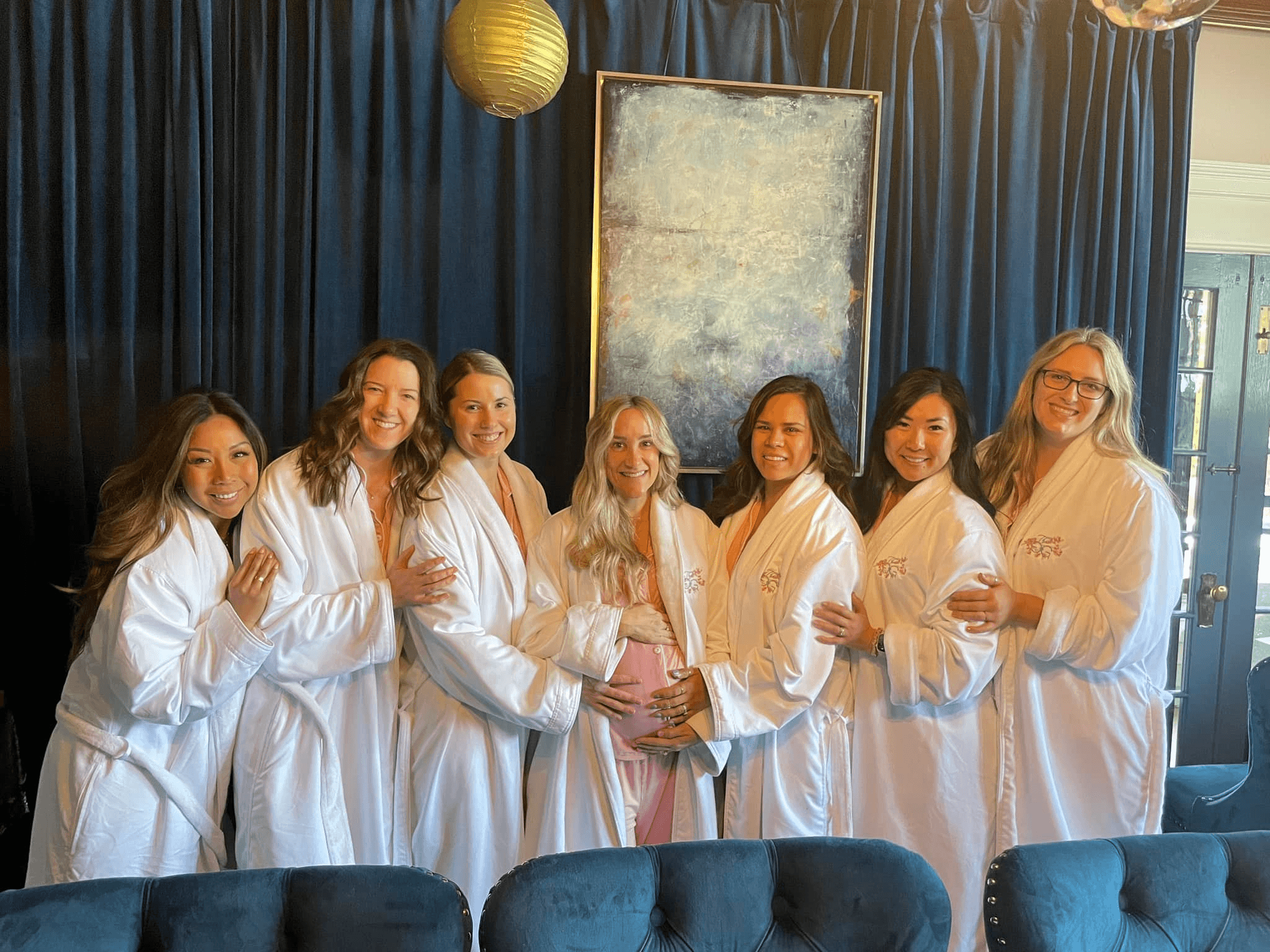 Seven ladies all wearing matching white bathrobes over pajamas standing in a row in front of a dark blue curtain