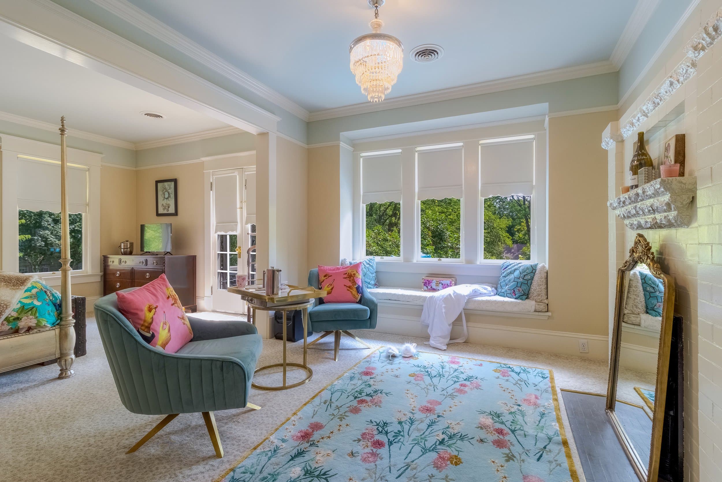 Two teal chairs set in the sitting area of a bedroom with a floral rug and bright windows