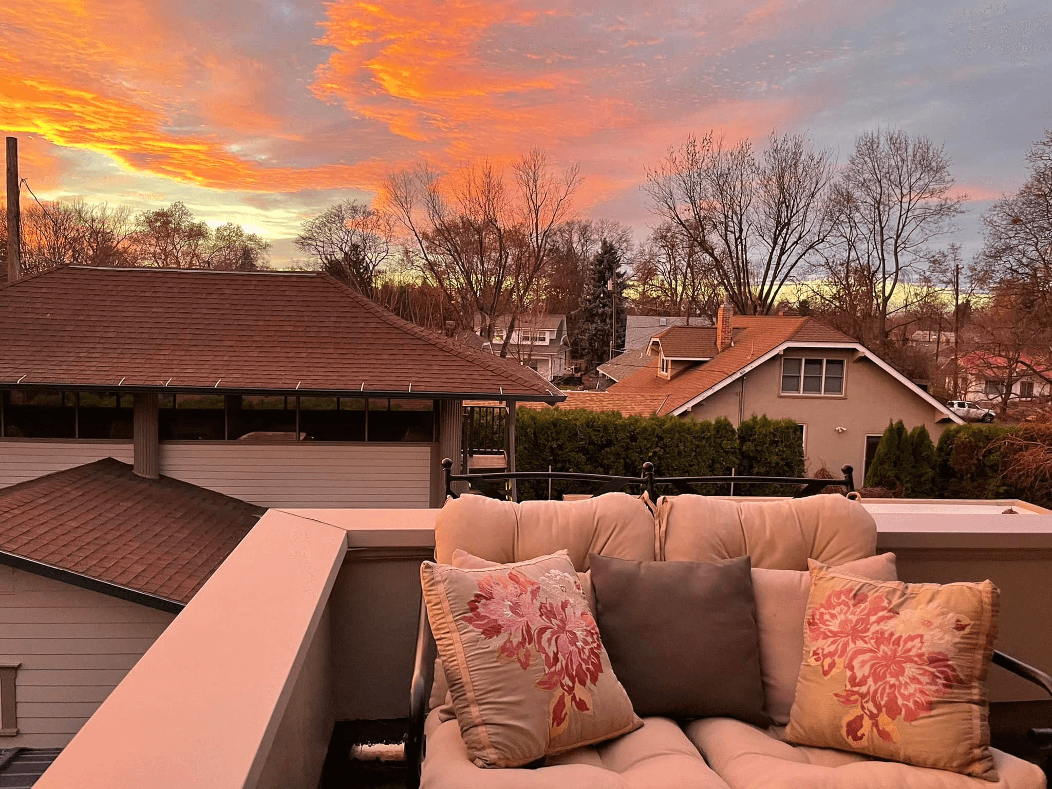 A cozy balcony view featuring a cushioned loveseat and a vibrant sunset sky over neighborhood rooftops.
