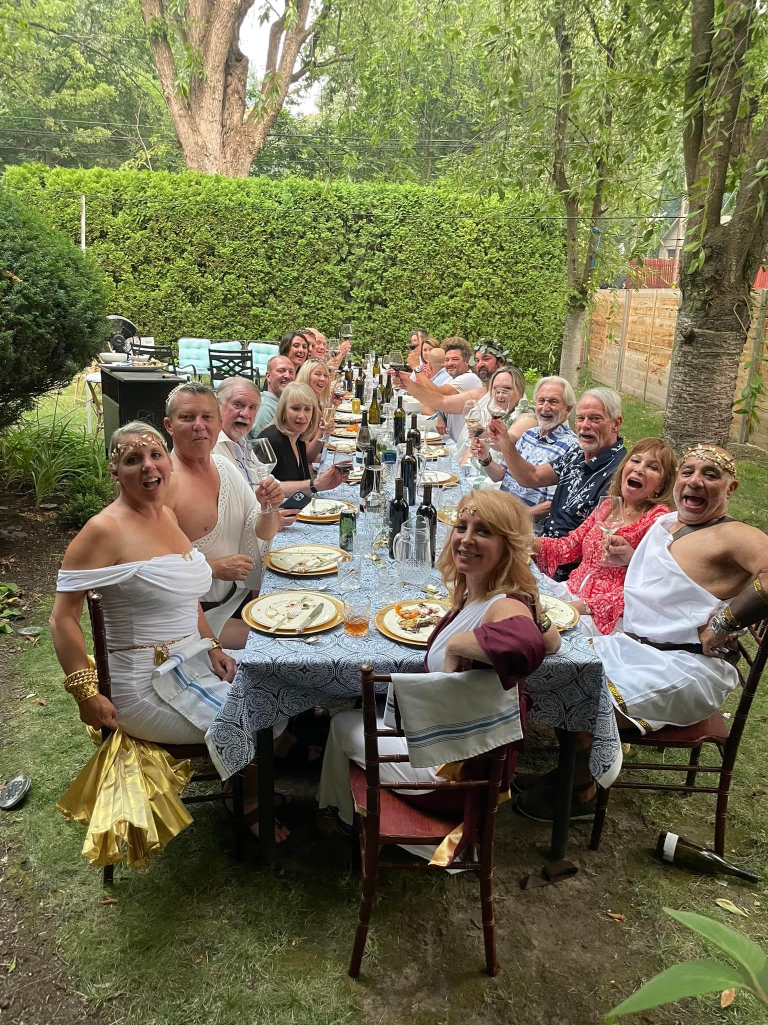A large group of people dressed in festive attire gather around a long table outdoors, celebrating with food and drinks.