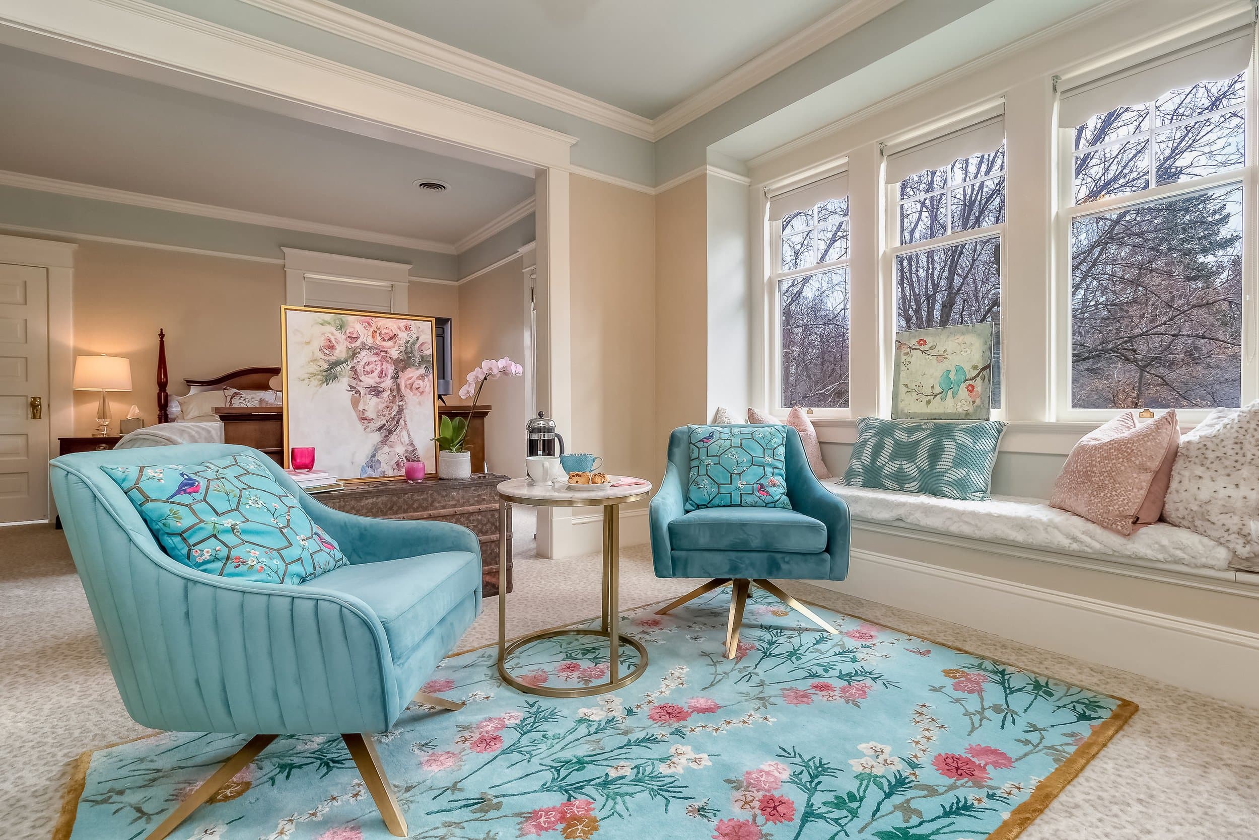 A cozy sitting area features two teal chairs, a marble-top table, and a colorful floral rug.