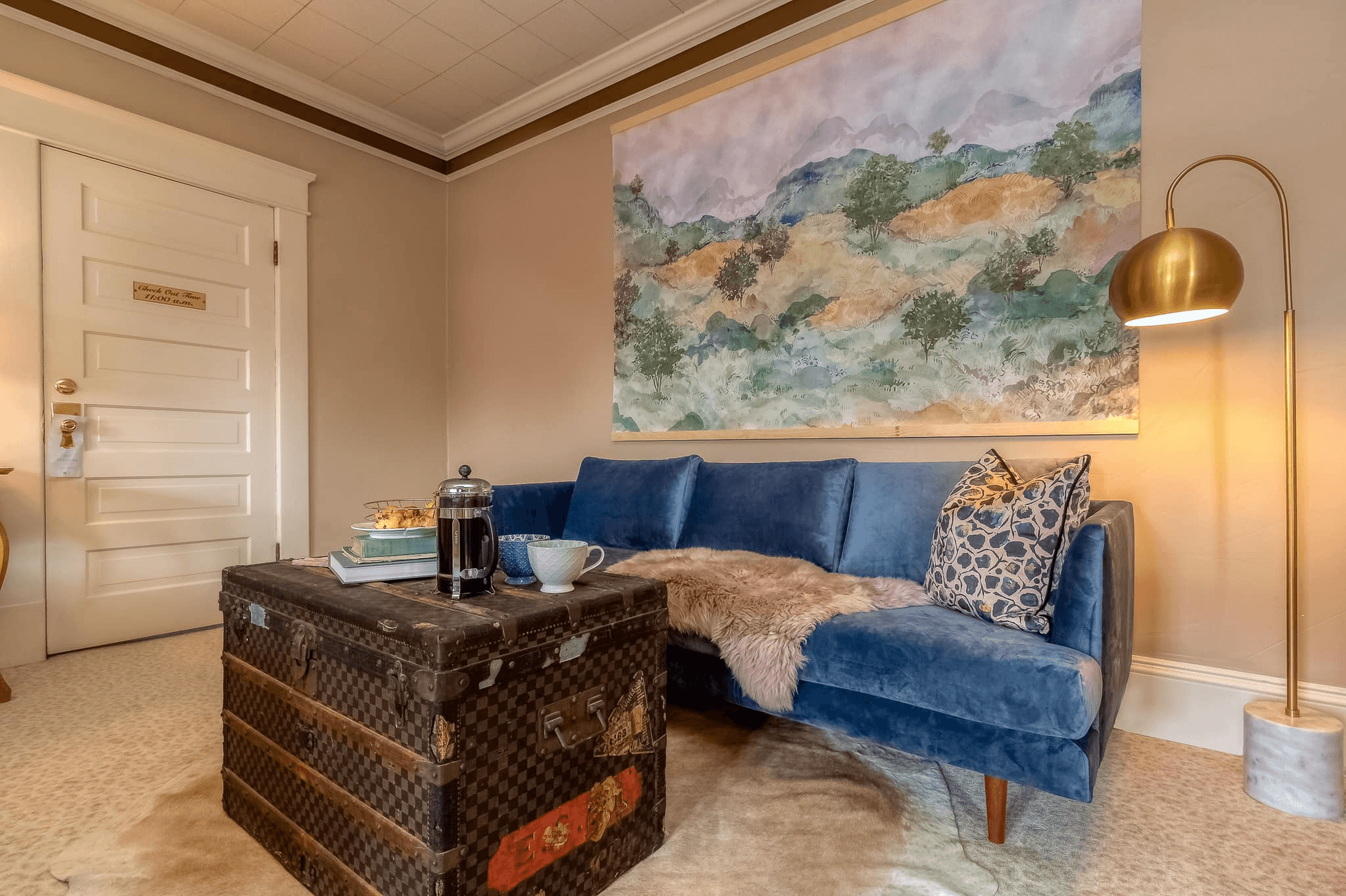 A cozy living space featuring a blue couch, a vintage trunk as a coffee table, and a large landscape painting on the wall.