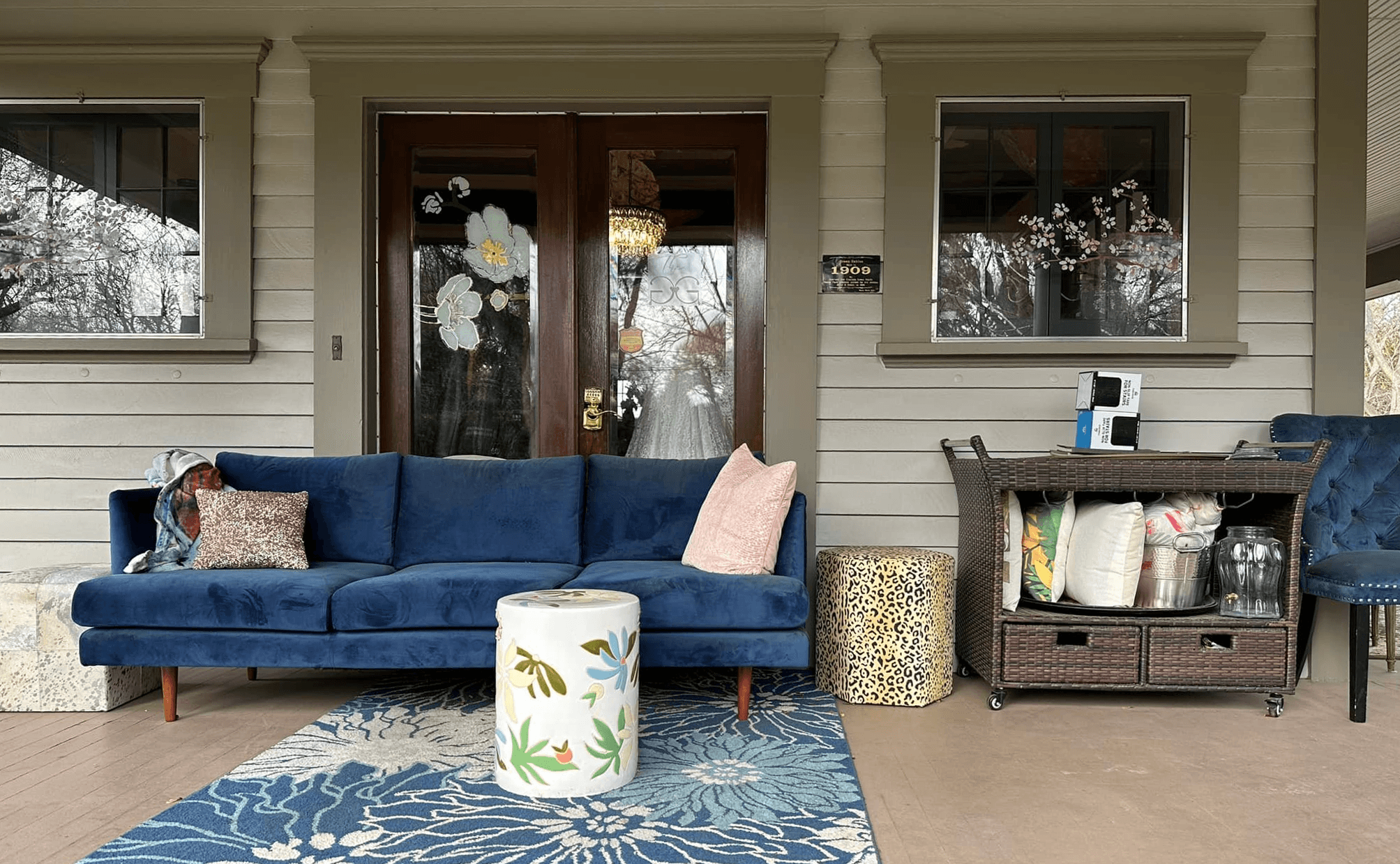 A cozy porch featuring a blue sofa, patterned rug, decorative side tables, and a storage cart.