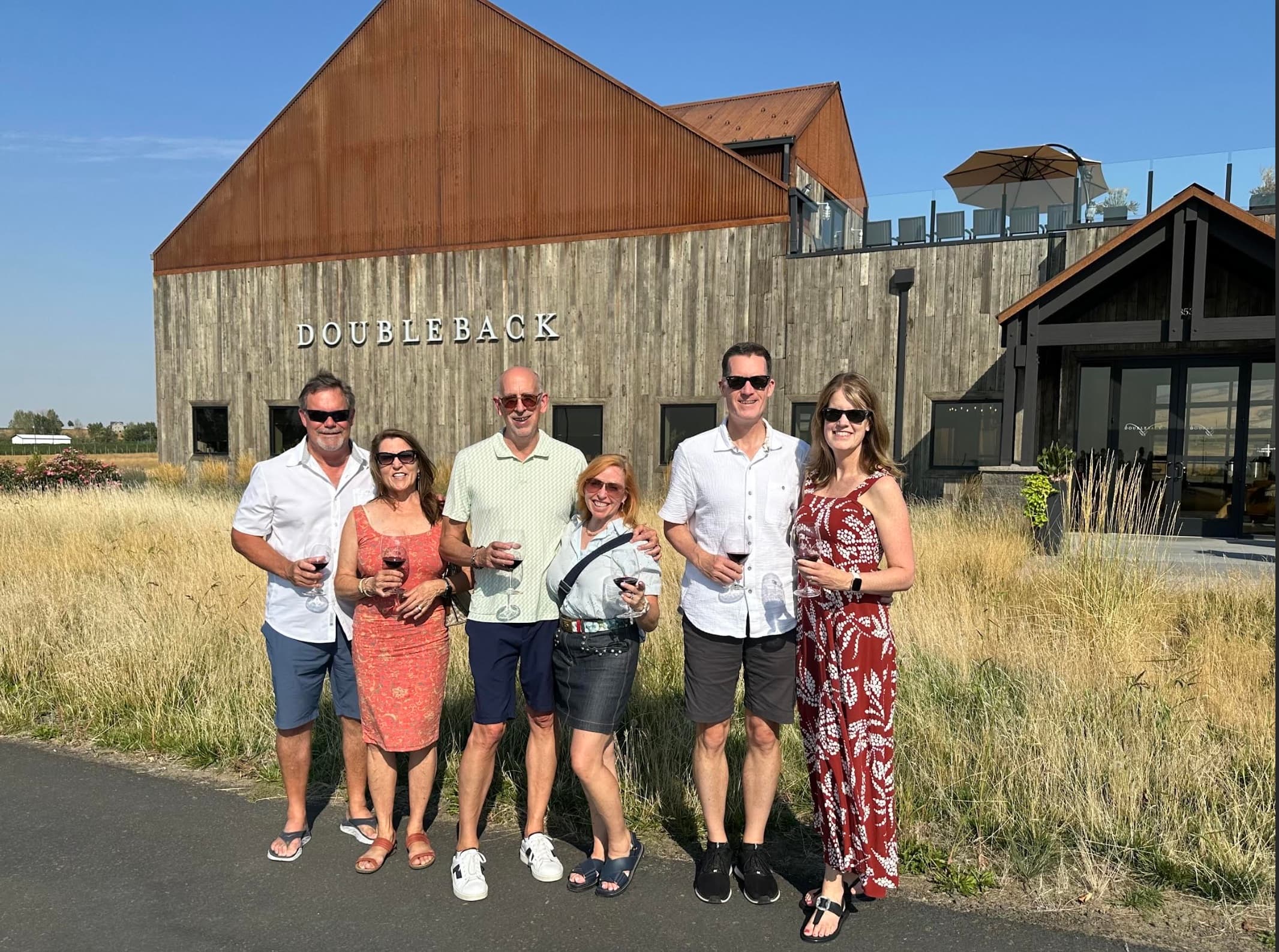 A group of six smiling guests holding glasses of red wine outside Doubleback Winery in Walla Walla, enjoying a sunny day during a curated retreat experience.